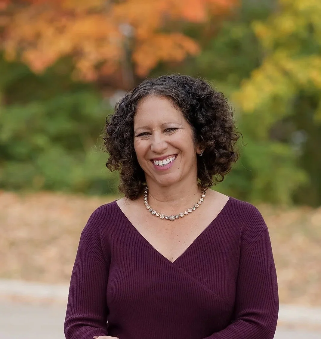 A woman with curly dark hair smiling outdoors in front of blurred autumn foliage, wearing a burgundy dress and a pearl necklace.