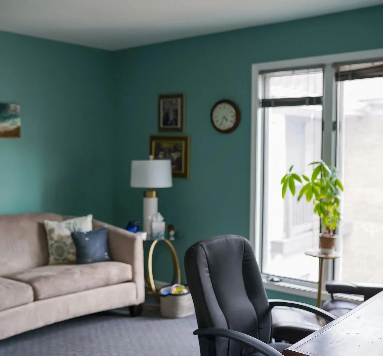 Therapy office with teal walls, beige sofa with pillows, a black office chair, a glass door leading outside, a potted plant on a stand, a table with chairs, a lamp on a side table, and framed pictures and a clock on the wall.