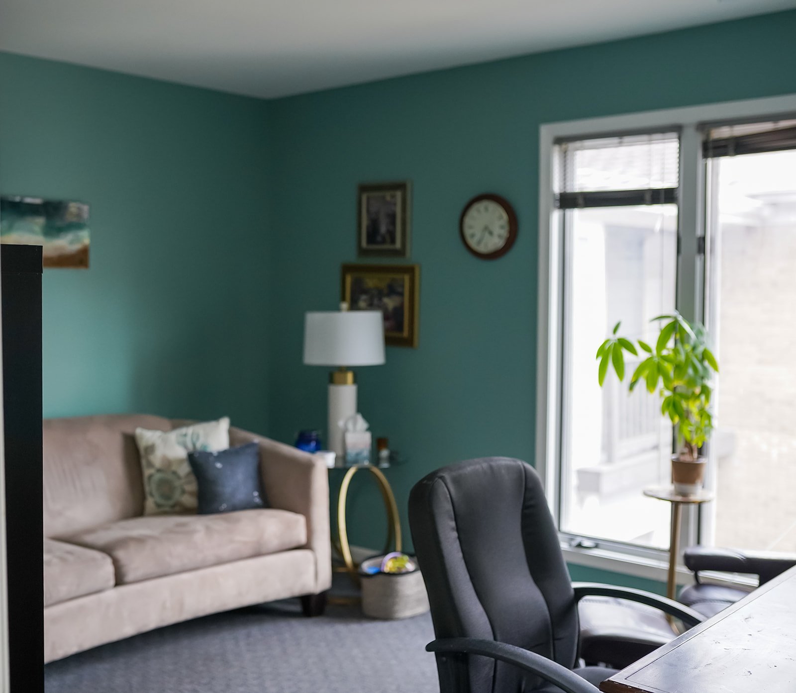 Therapy office with beige couch, black office chair, green wall, framed pictures, table lamp, large window with blinds, potted plant, and a clock on the wall.