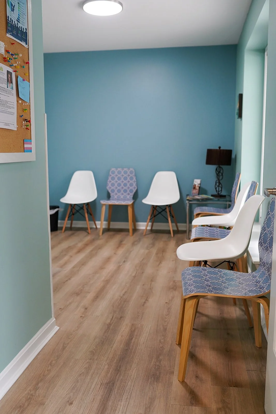 A waiting room with light wood flooring, three white chairs and three chairs with blue patterned fabric, a blue accent wall, a side table with magazines, a dark brown lamp, and a bulletin board with pinned notices.