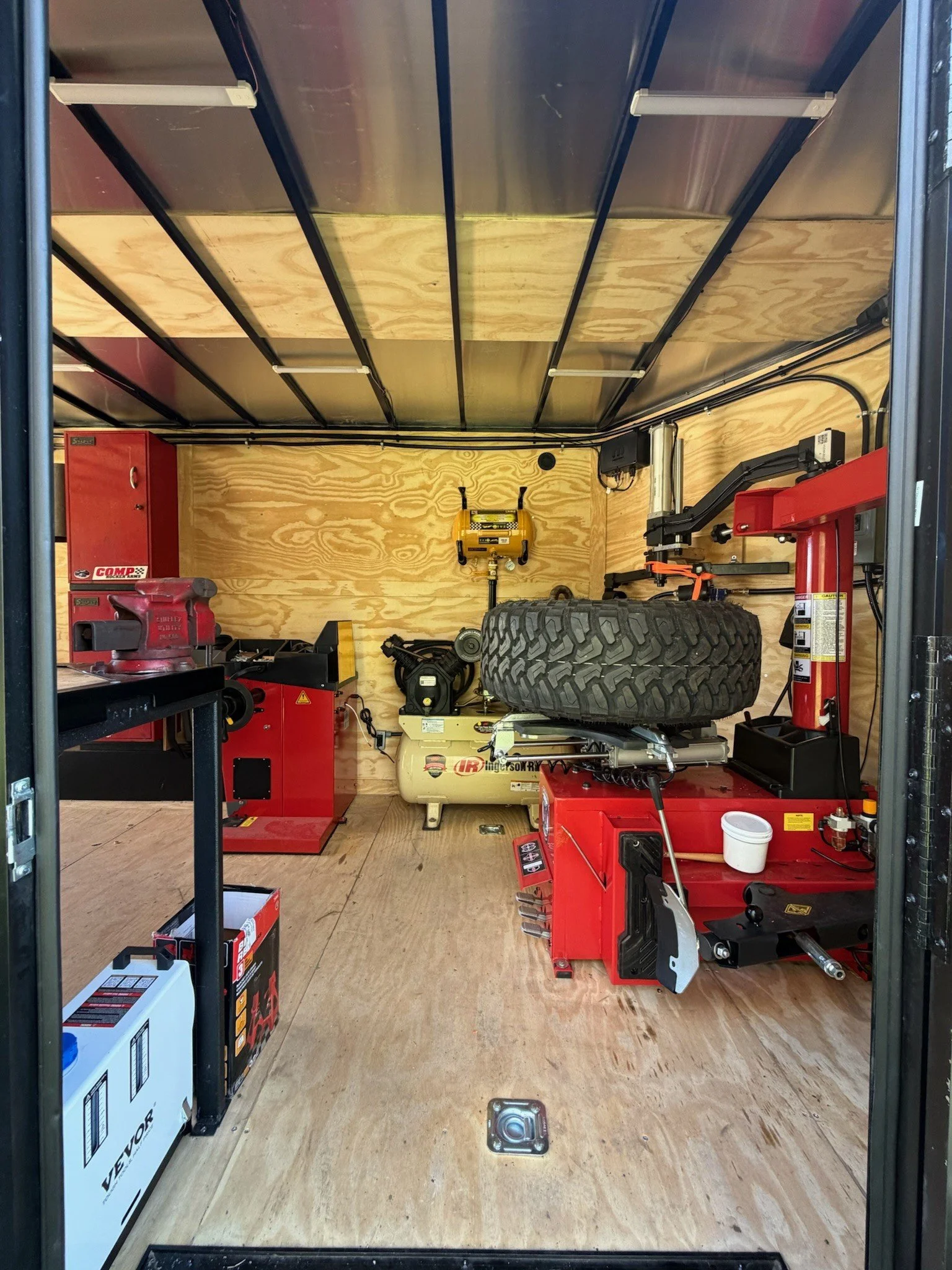 View of a small workshop or garage with wooden floors and plywood walls, containing various tools, equipment, and a larger tire, with a workbench on the left side and a tire changer on the right side.