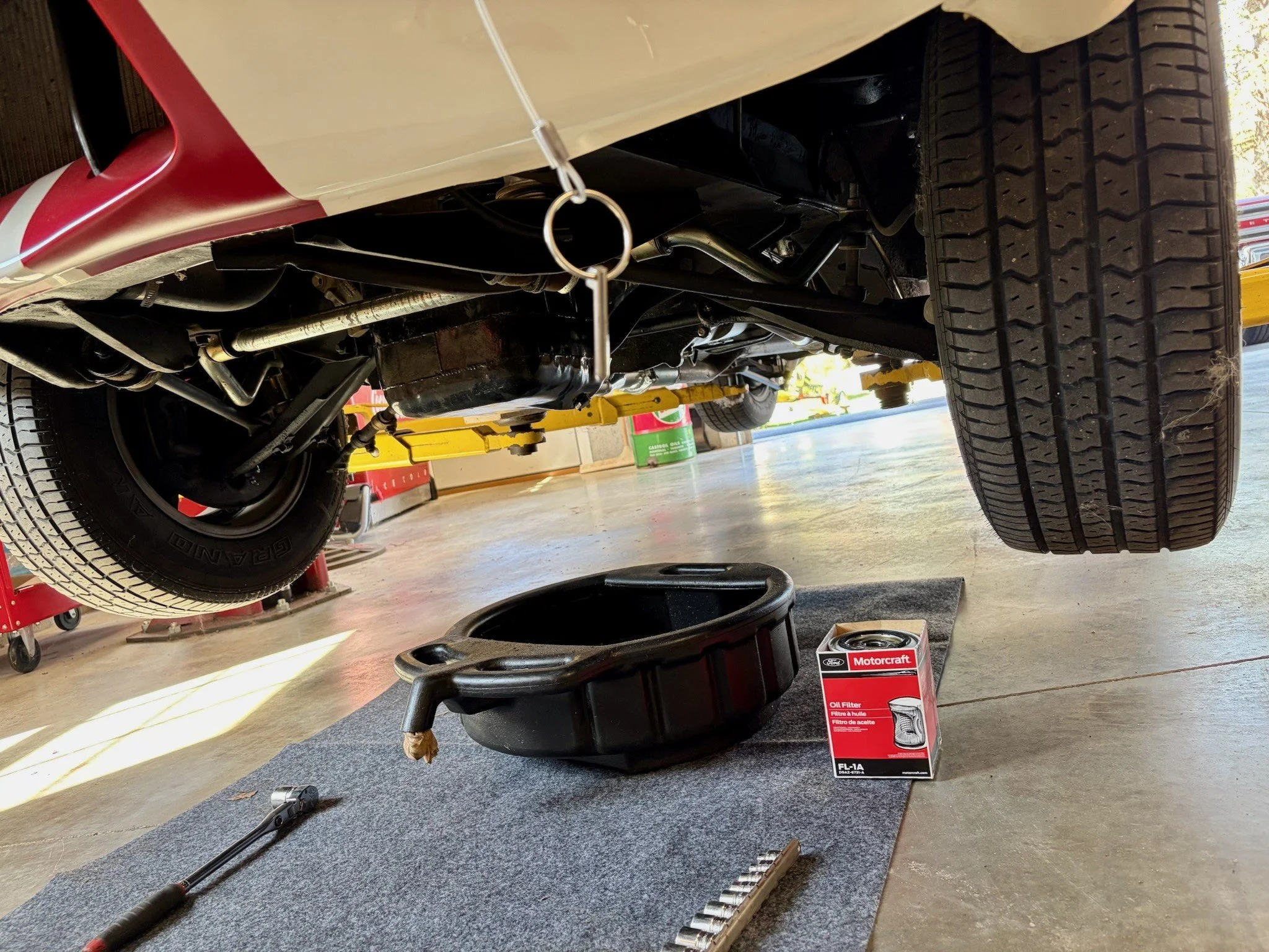 Underneath view of a vehicle lifted on a hydraulic lift, with a tire visible on the right side. Various automotive tools and a Motorcraft oil filter box are on the floor nearby.