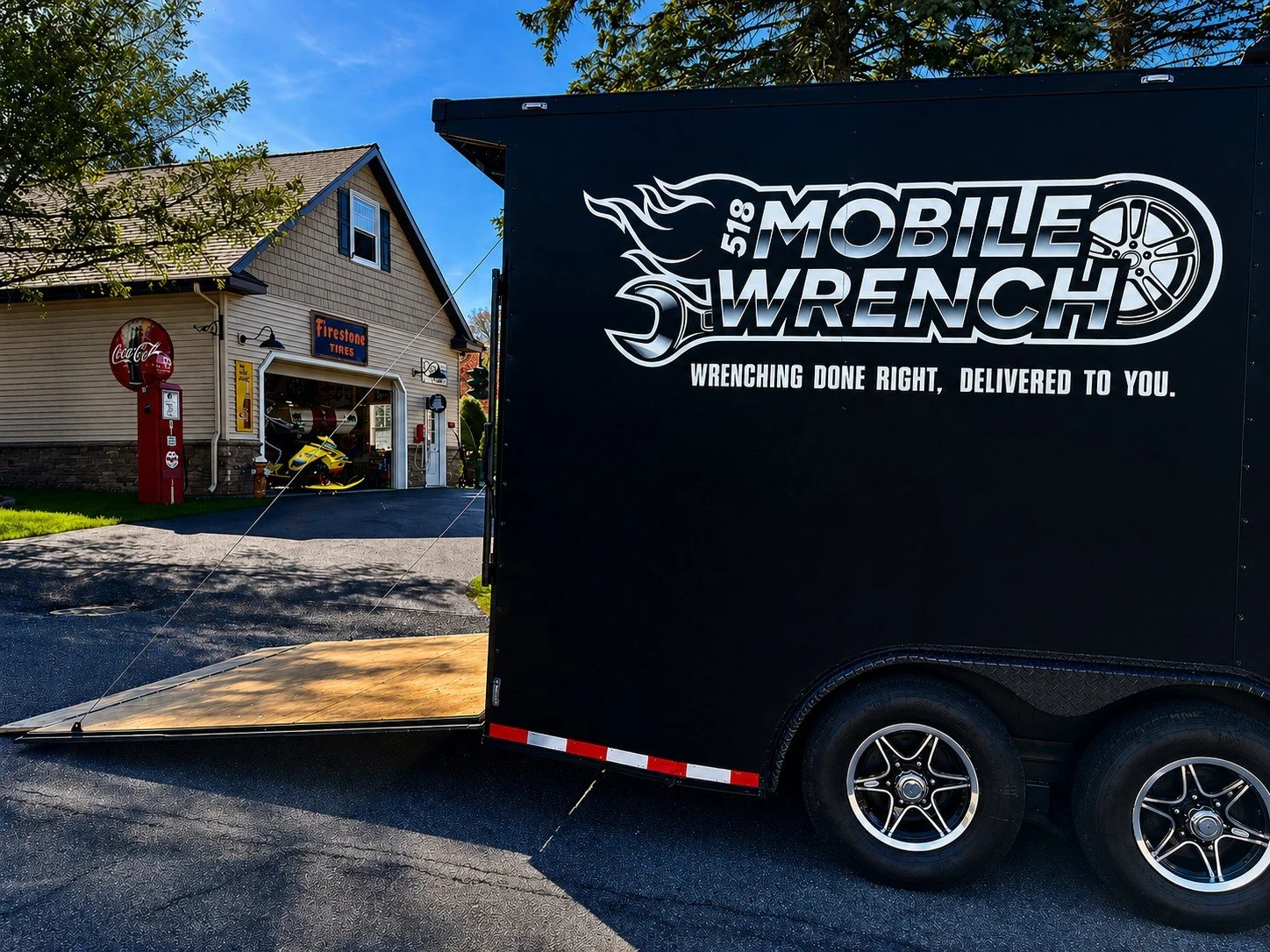 A mobile welding trailer parked on a driveway with a house in the background. The trailer has the branding 'Mobile Wrench' and 'Wrenching done right, delivered to you,' with a large wheel graphic. The house has a Firestone Tires sign, a red Coca-Cola vending machine, and a garage with a yellow ATV inside.