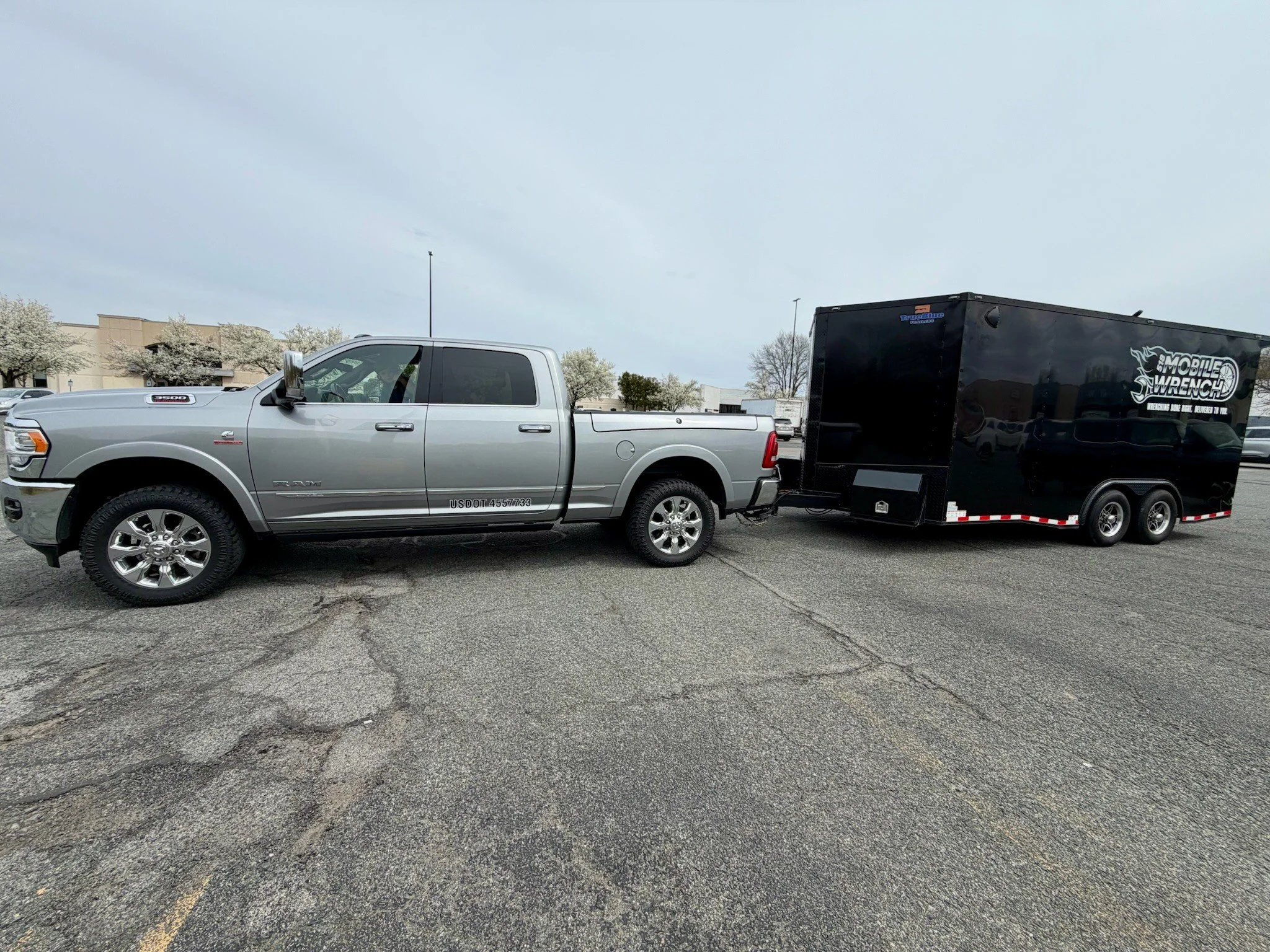 A silver Ram pickup truck towing a black mobile wrench trailer in a parking lot with a building and flowering trees in the background.