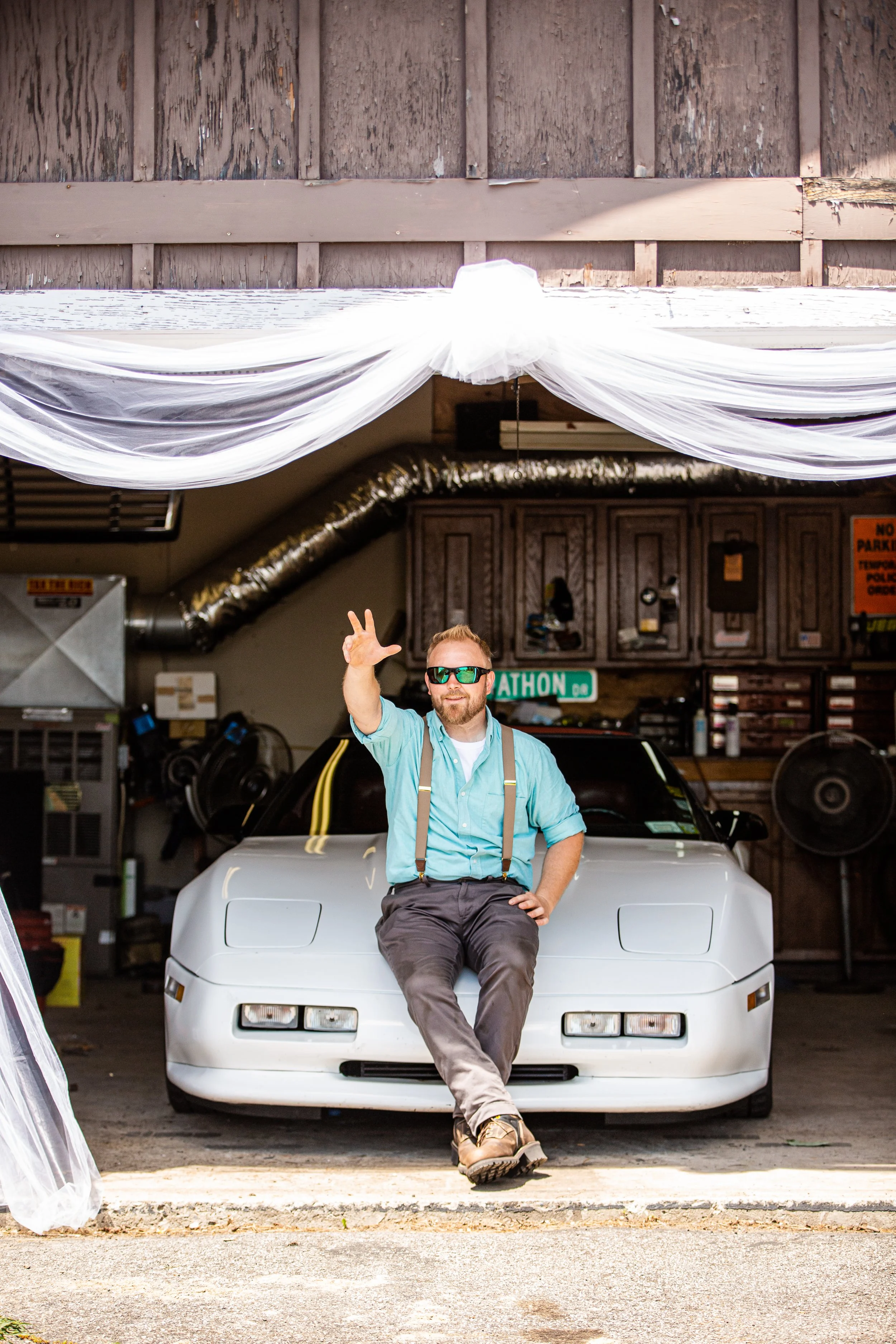 A man with sunglasses and a beard, sitting on the hood of a white sports car, waving with one hand inside a garage with wood-paneled walls and various tools and equipment.