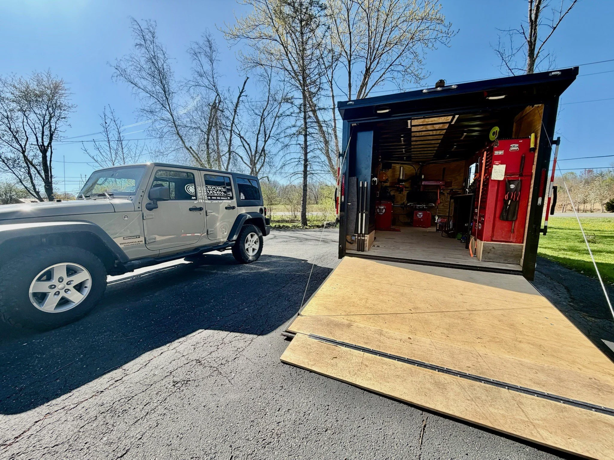 A silver Jeep parked next to an open trailer filled with tools and equipment, including a red toolbox and various hand tools, in an outdoor area with trees and a clear blue sky in the background.