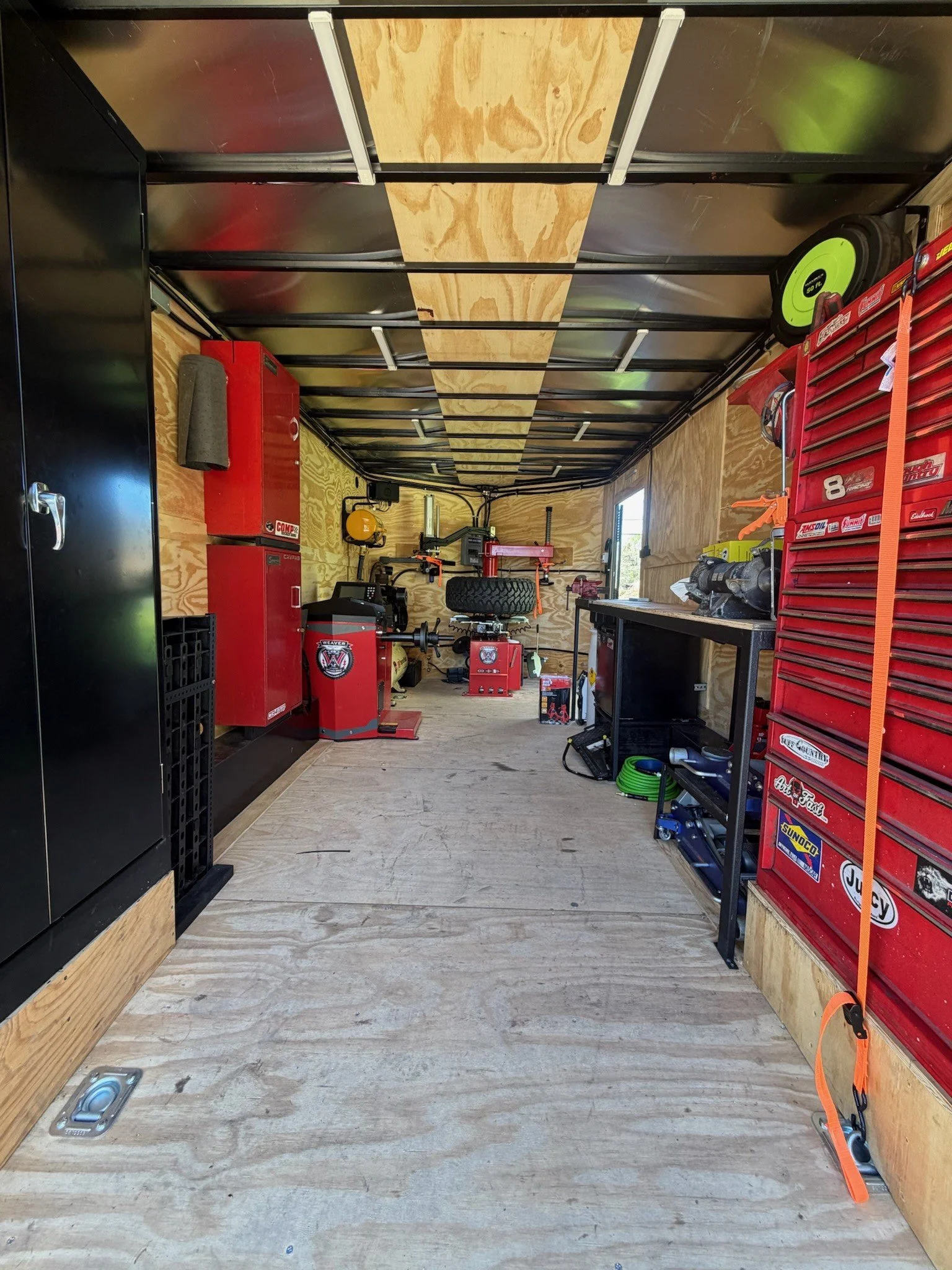 The interior of a workshop or garage with wooden walls and floor. The space contains red tool chests, a black workbench, and various tools and equipment, including a yellow extension cord reel, a tire, and power tools. There is a window on the right wall and a ceiling frame with some metal and wooden support.