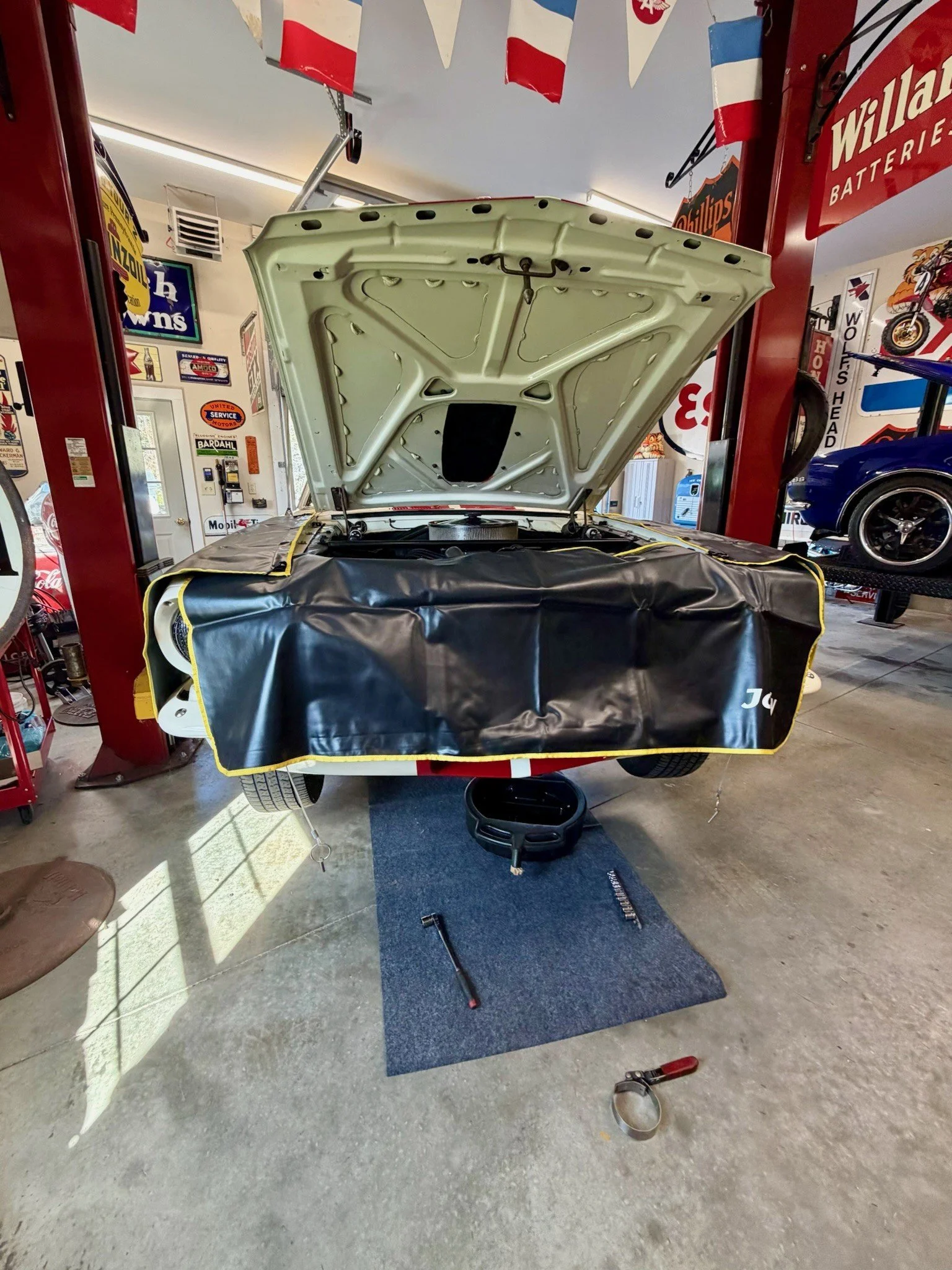 A car in a garage with its hood open and the front end lifted on a hydraulic lift. The garage has automotive memorabilia and signs on the walls, and there are tools and equipment on the floor.