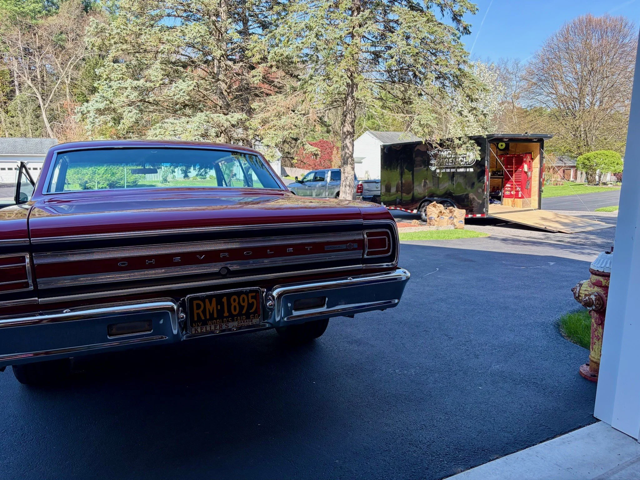A vintage red Chevrolet car parked in a driveway, with a black trailer behind it, surrounded by trees and a suburban neighborhood under a clear blue sky.