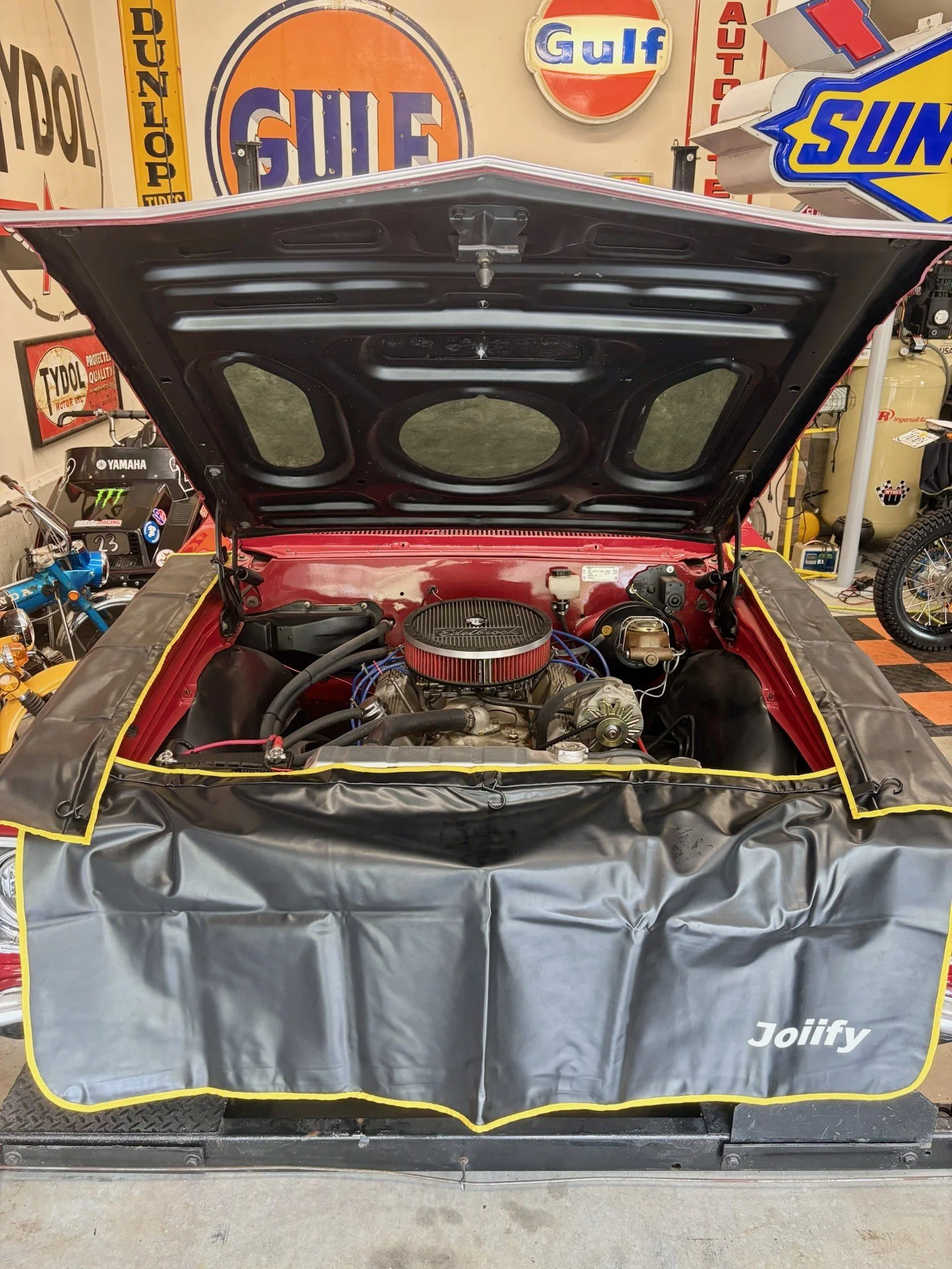 Open car hood showing engine with red air filter, surrounded by car garage items including signs, a bicycle, and a tire.