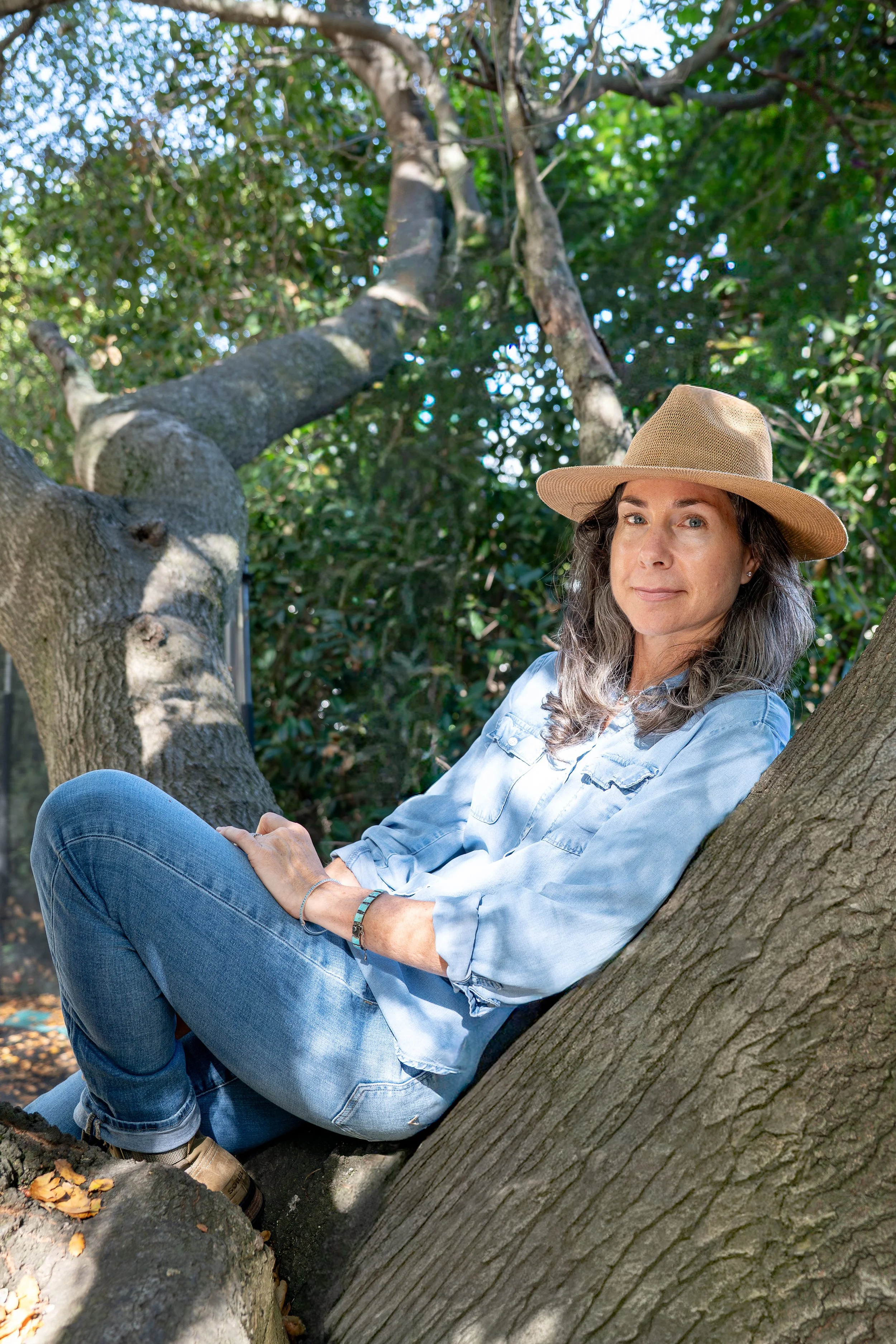 A woman sitting in a tree, wearing a beige hat and a light blue shirt, surrounded by green foliage.