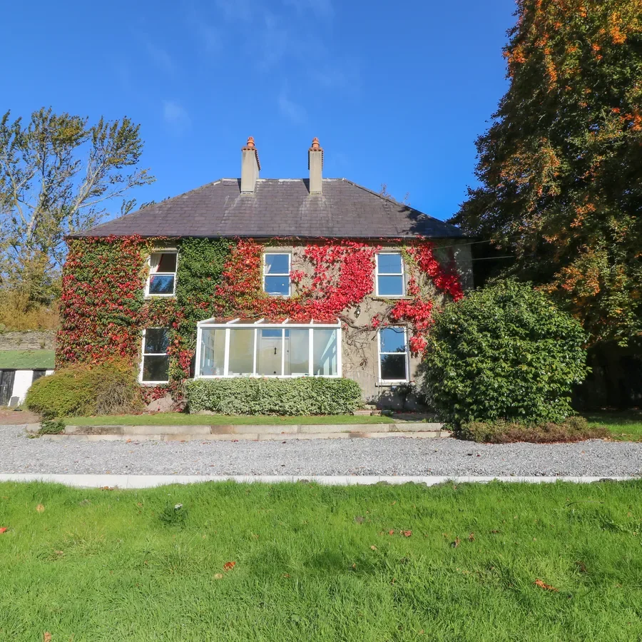 A house with a gray stone exterior partially covered in green and red ivy, with four windows and a glass-enclosed porch, surrounded by trees and a well-maintained lawn under a bright blue sky.