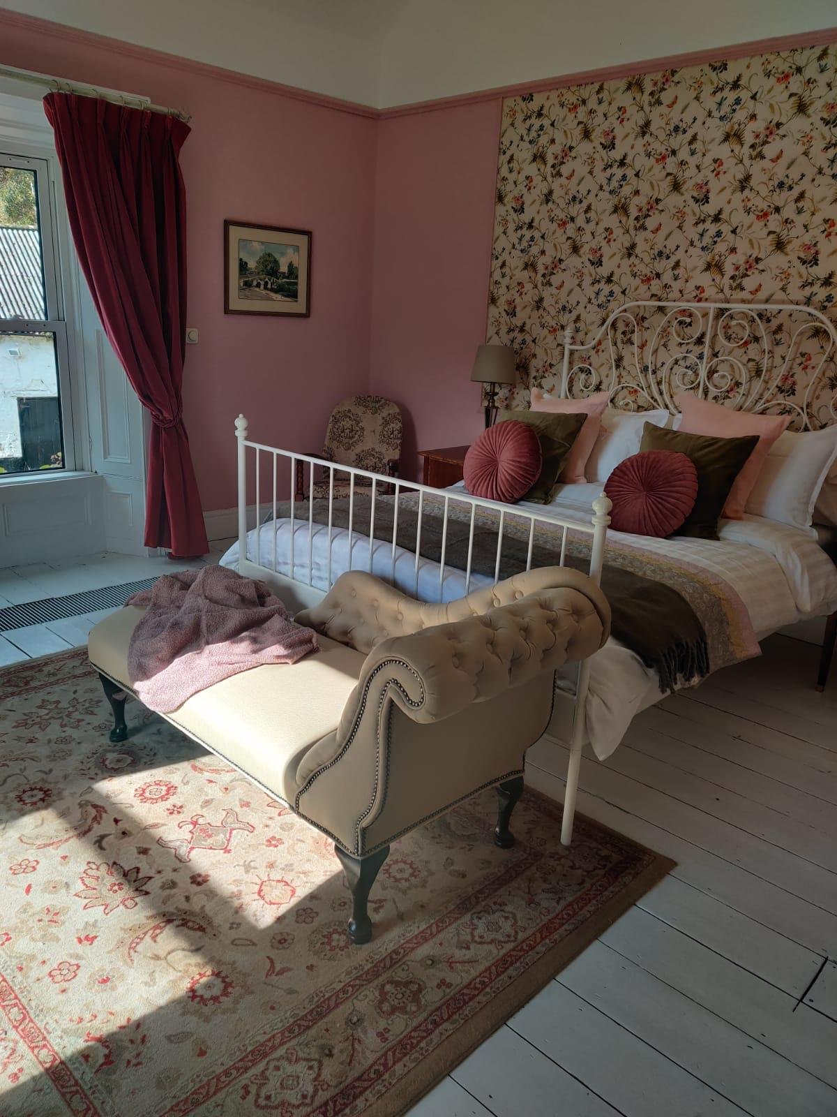 A cozy bedroom with pink walls, a white metal bedframe, floral wallpaper headboard, and a variety of pillows including round pink and square green ones. There is a beige tufted chaise lounge at the foot of the bed with a pink blanket. A stone upholstered chair, a wooden nightstand with a lamp, and a window with pink curtains are also visible. An area rug with floral patterns is on the floor.