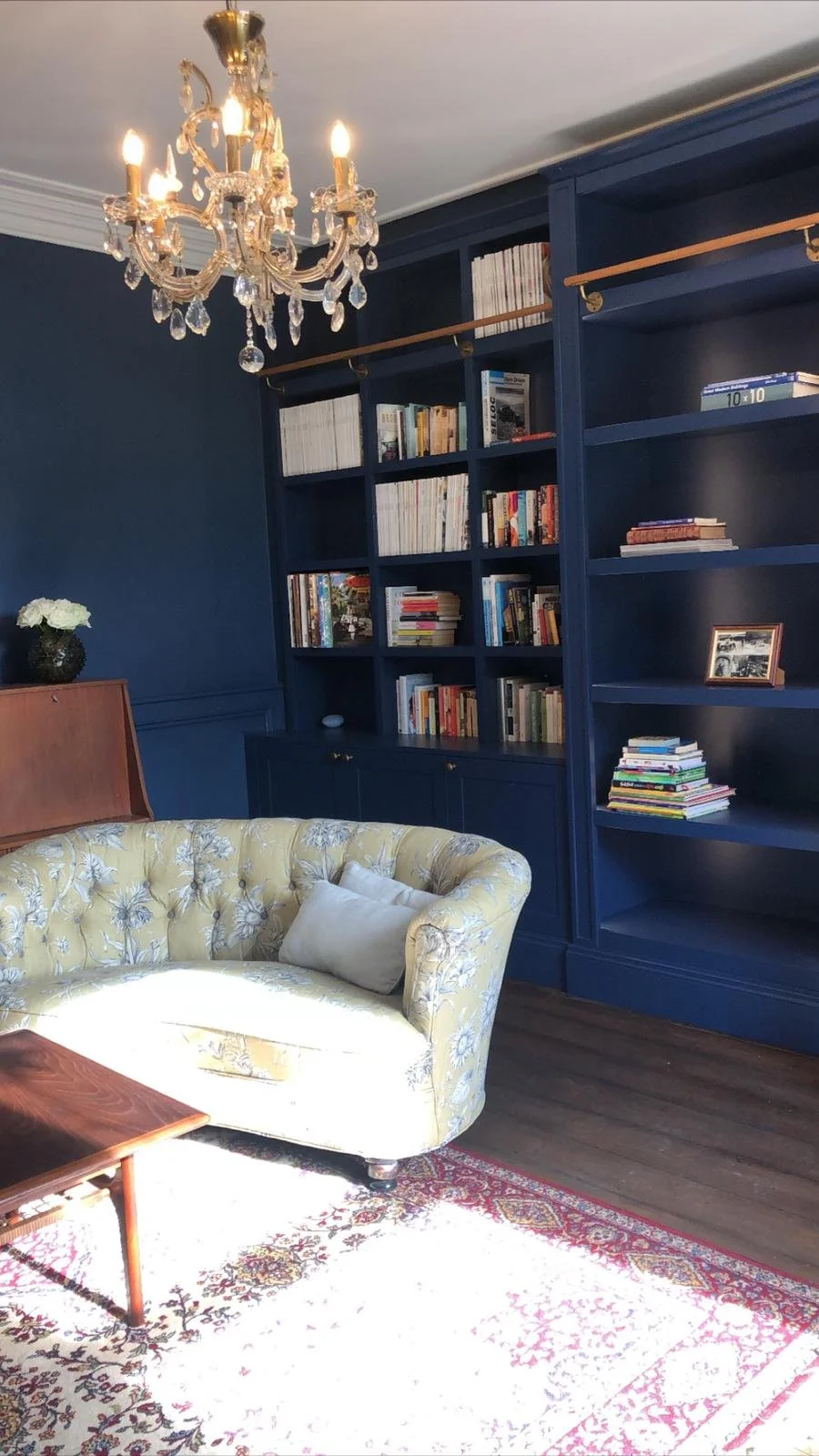 A living room with a yellow floral patterned sofa, a dark blue bookshelf filled with books, a chandelier hanging from the ceiling, and a vintage wooden coffee table on a white and red patterned rug.