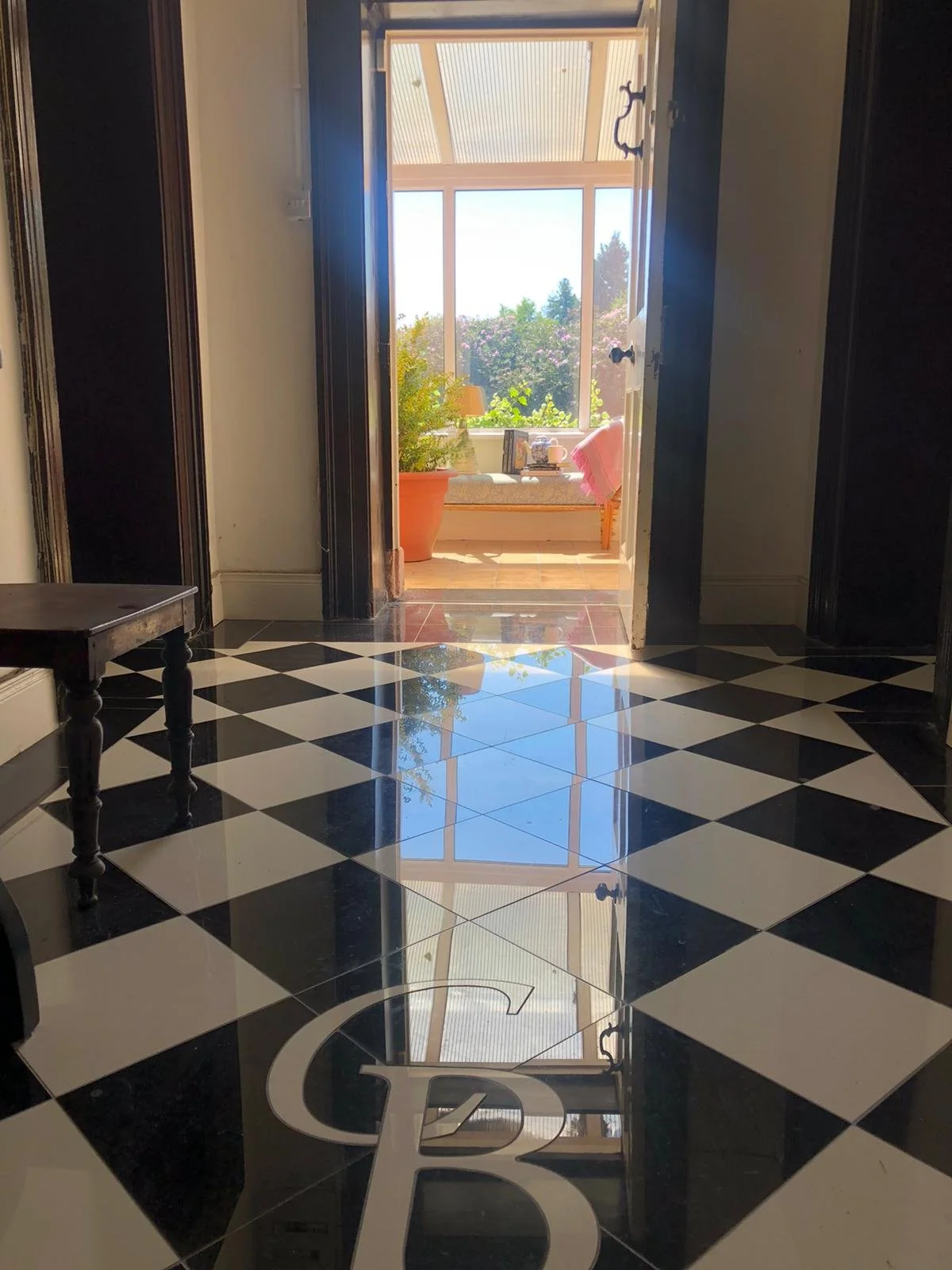 View of a sunlit conservatory or porch with large windows, potted plants, a small table, and a pink towel or cloth draped over a chair, seen from an entryway with black and white checkered floor reflected on the glossy surface.