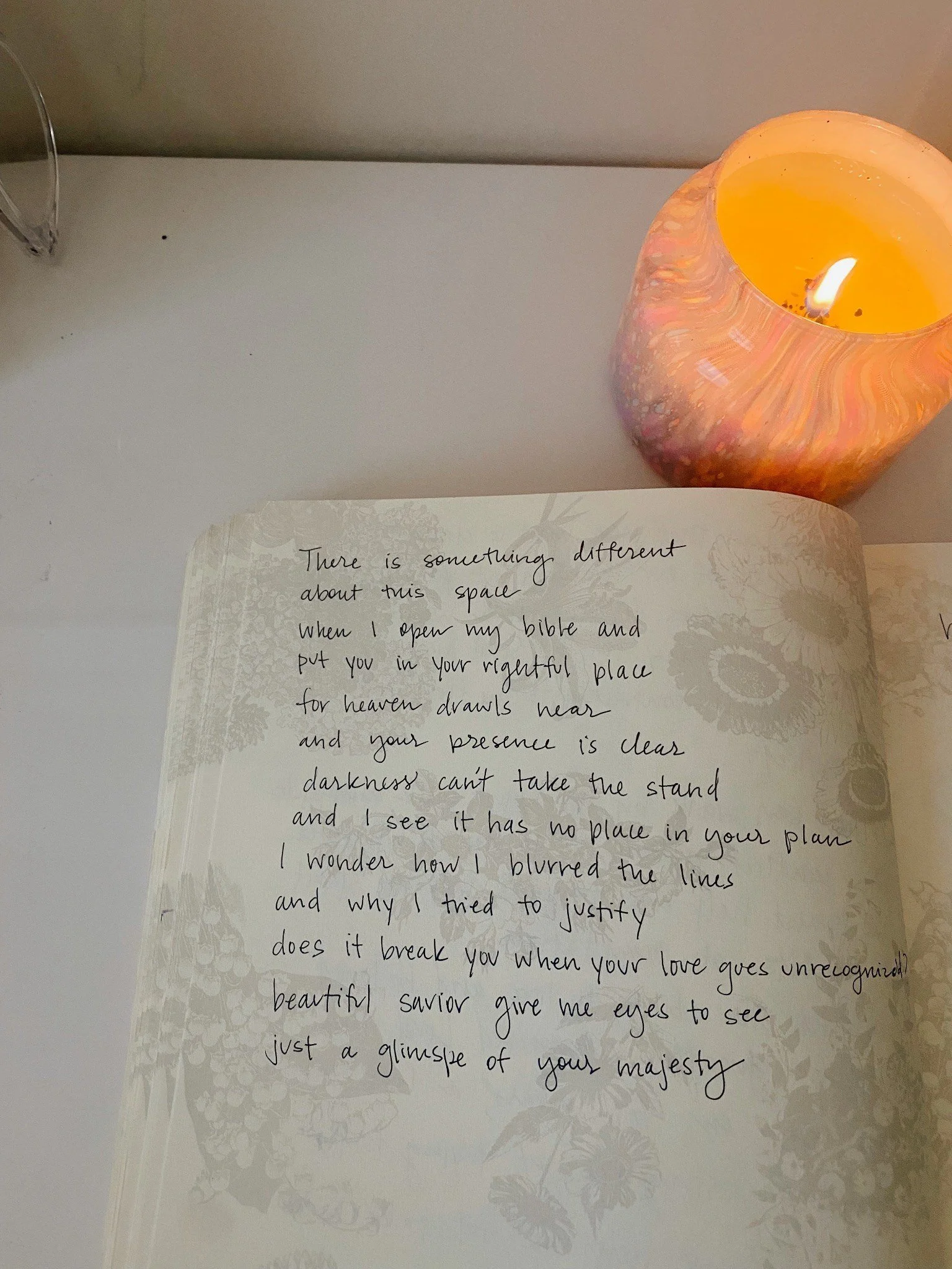 A notebook with handwritten poetic text, a lit candle in a pink and orange marbled holder, on a white table.