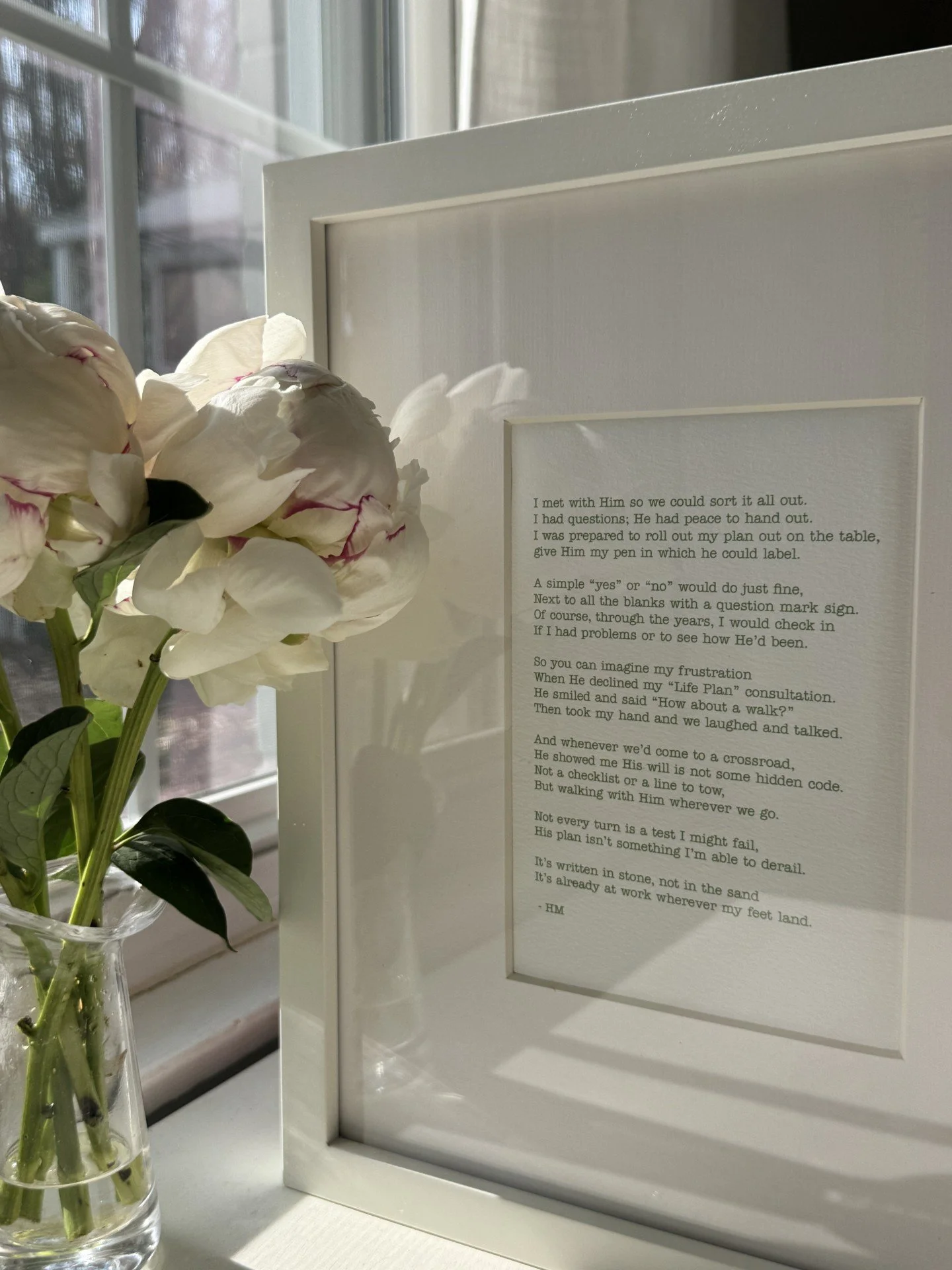 A framed poem on a white wall, with a vase of white flowers next to it, placed on a windowsill with sunlight coming through the window.