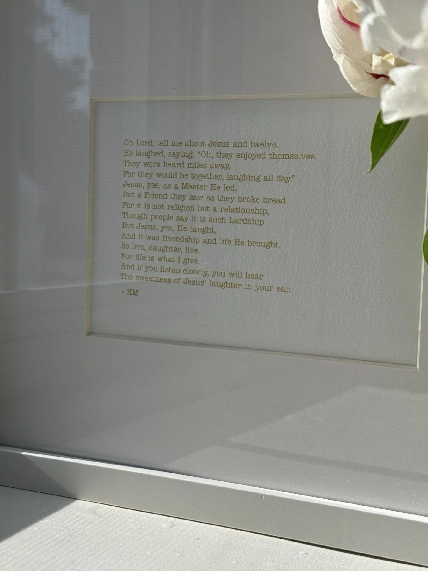 A framed poem titled 'Oh Lord, tell me about Jesus and twelve' with gold text on a white background, partially covered by white flowers with pink accents, sitting on a white surface.