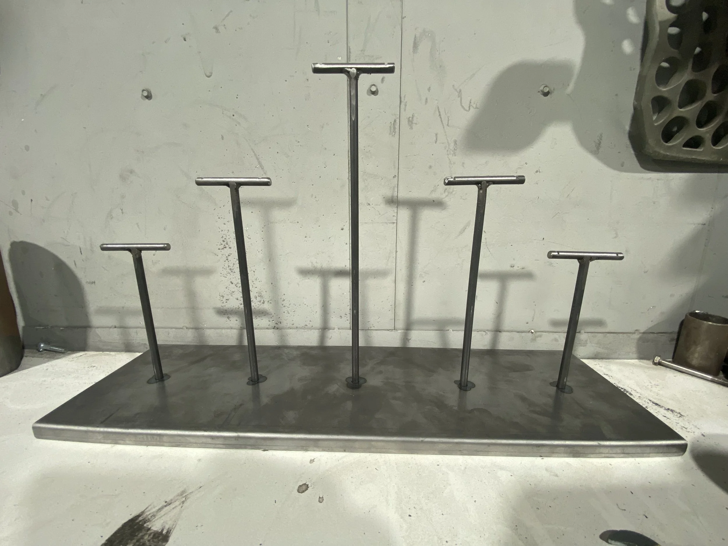 Set of metal T-shaped objects standing upright on a metal base, arranged in a row, with shadows cast on a white wall behind them.