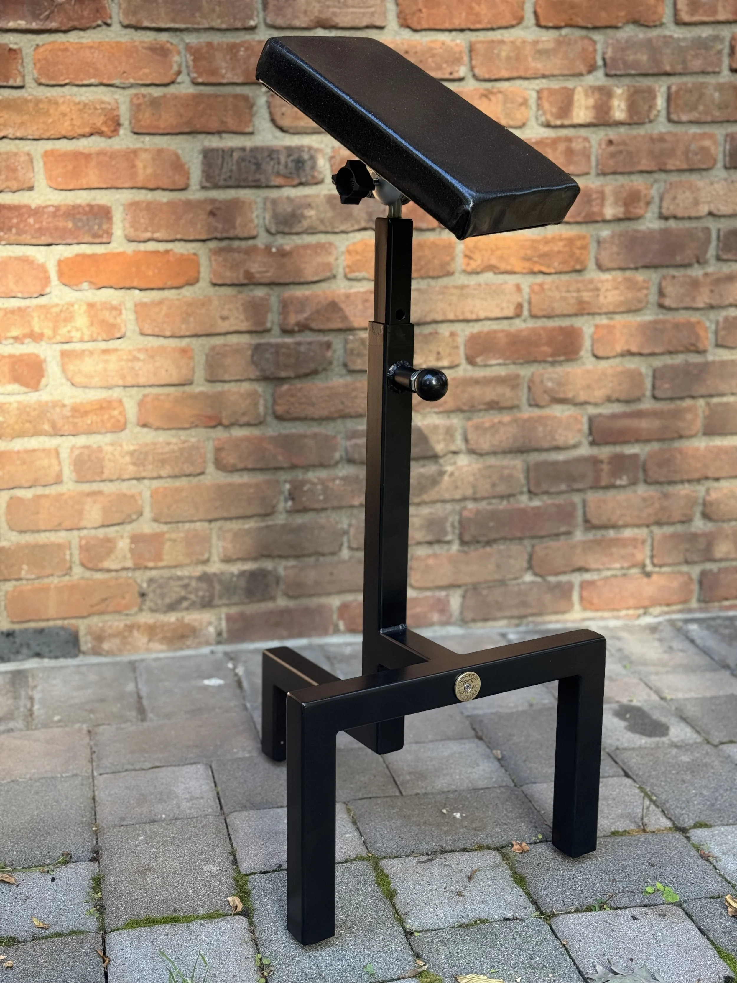 A black adjustable footstool with a cushioned top, positioned on a paved brick surface against a brick wall background.