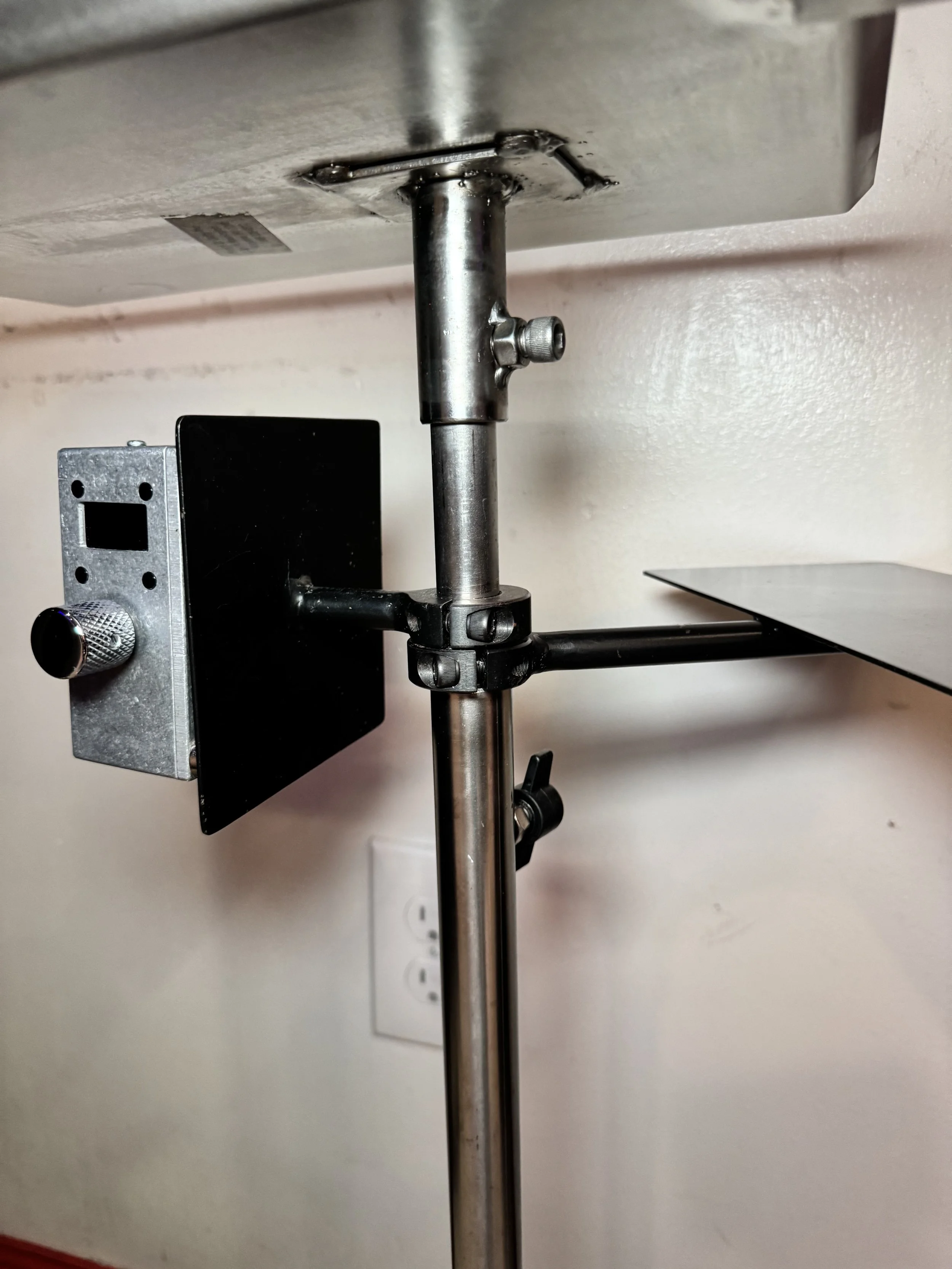 Close-up view of a tripod stand with adjustable knobs, mounted underneath a table or counter, with a black box attached to the side, and a white electrical outlet on a cream-colored wall in the background.