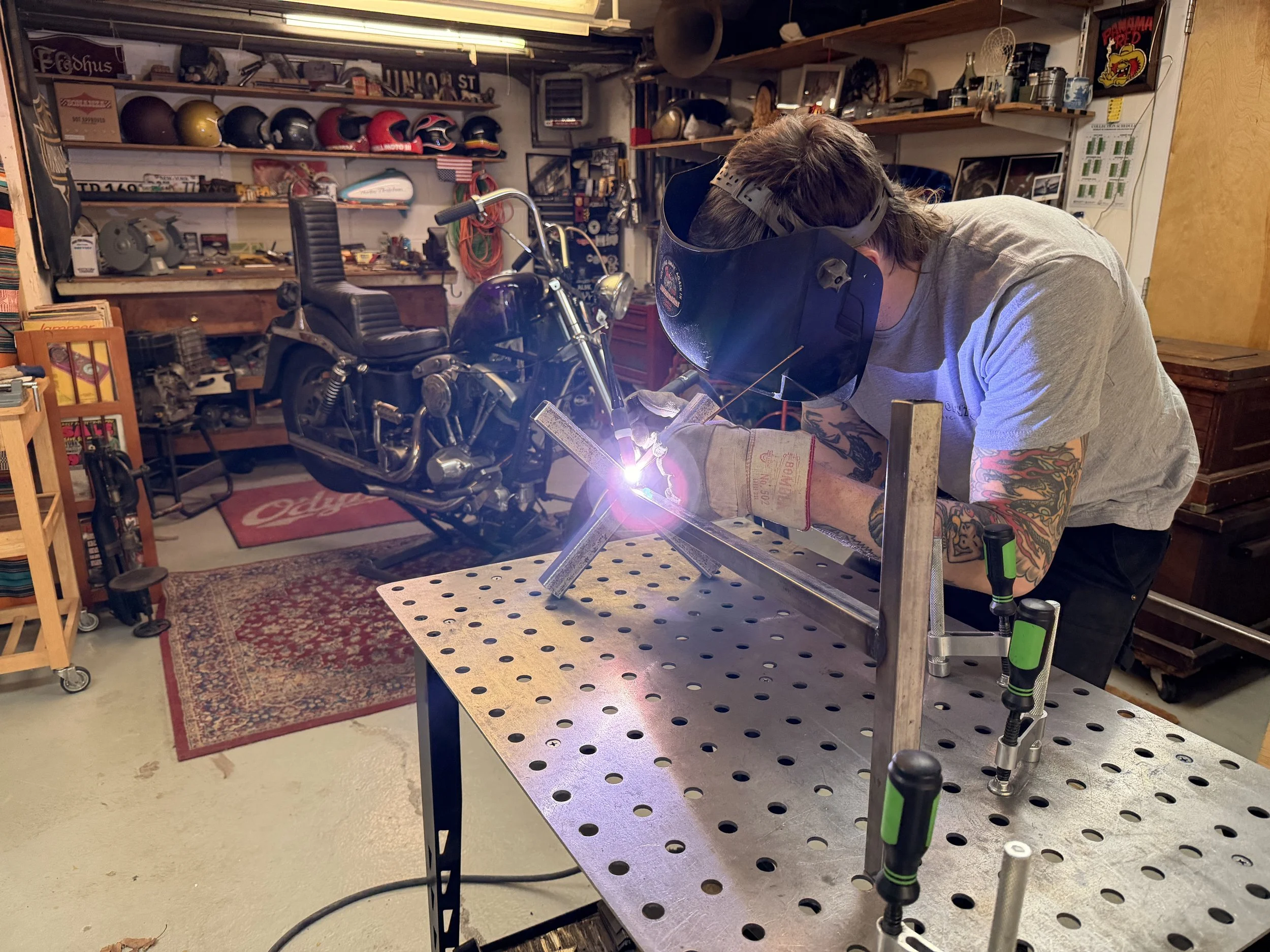 A person welding a metal frame in a garage workshop, with a motorcycle and various tools in the background.