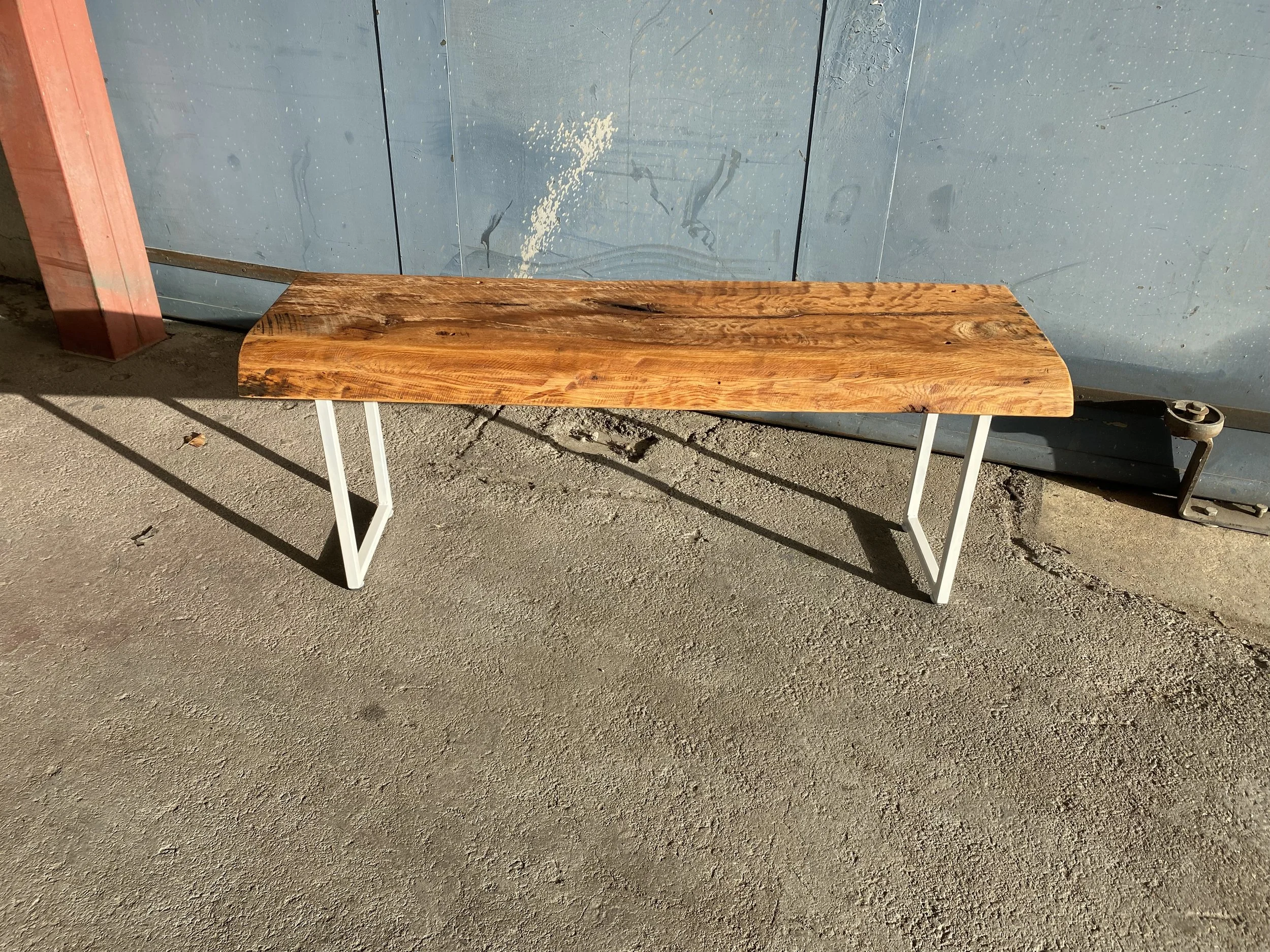 A wooden bench with white metal legs on a concrete surface with a blue wall in the background.