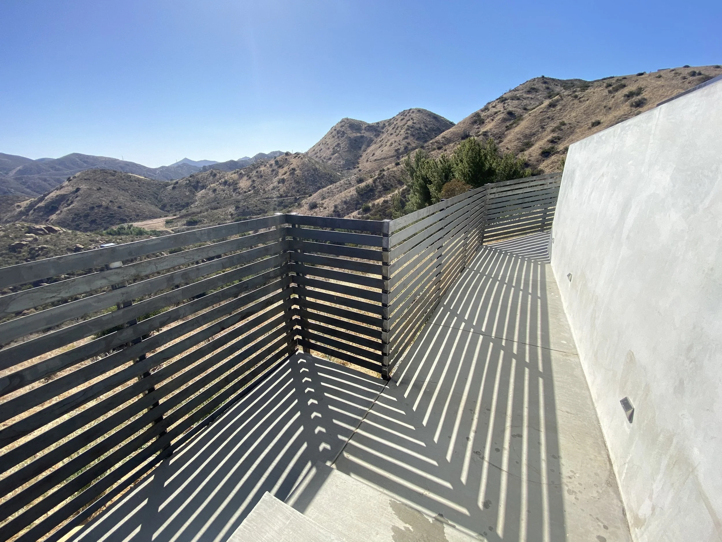 View of a balcony with metal railing casting striped shadows, overlooking dry rolling hills under a clear blue sky.