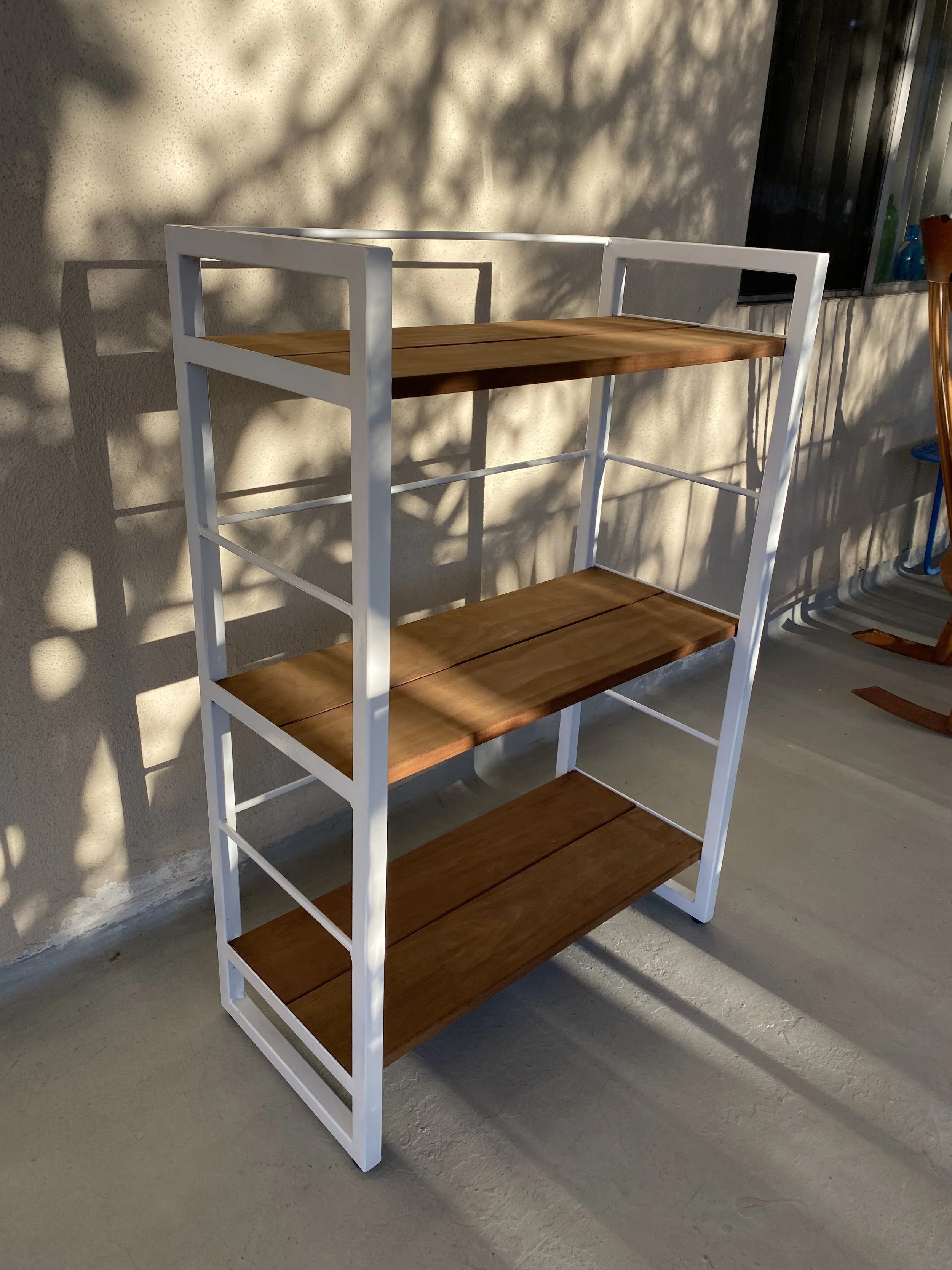 A multi-level wooden and white metal bookshelf standing on a concrete floor, with shadows cast on the wall behind it.