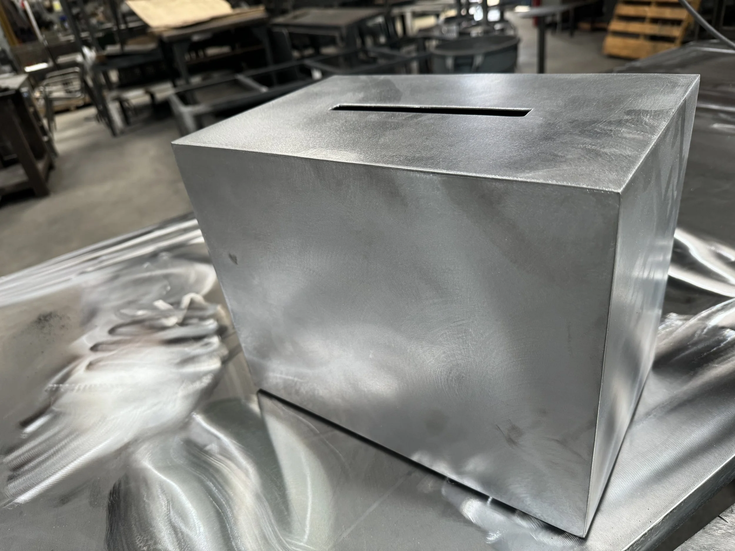 A metallic rectangular box with a slot on the top, placed on a reflective metal surface in an industrial setting.