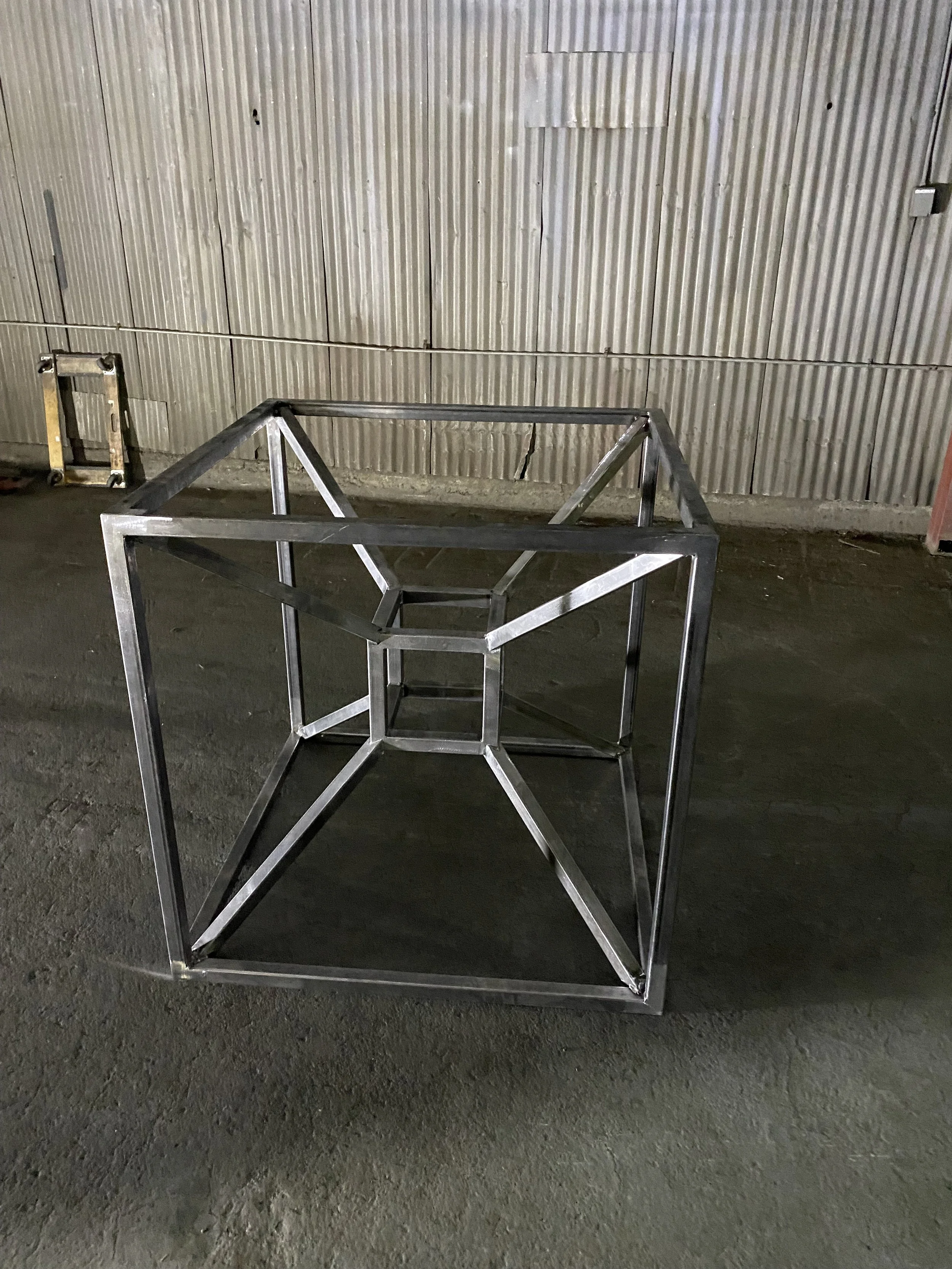 Metal frame structure resembling a cuboid shape with a star-shaped opening in the middle, set against a corrugated metal wall and a concrete floor.
