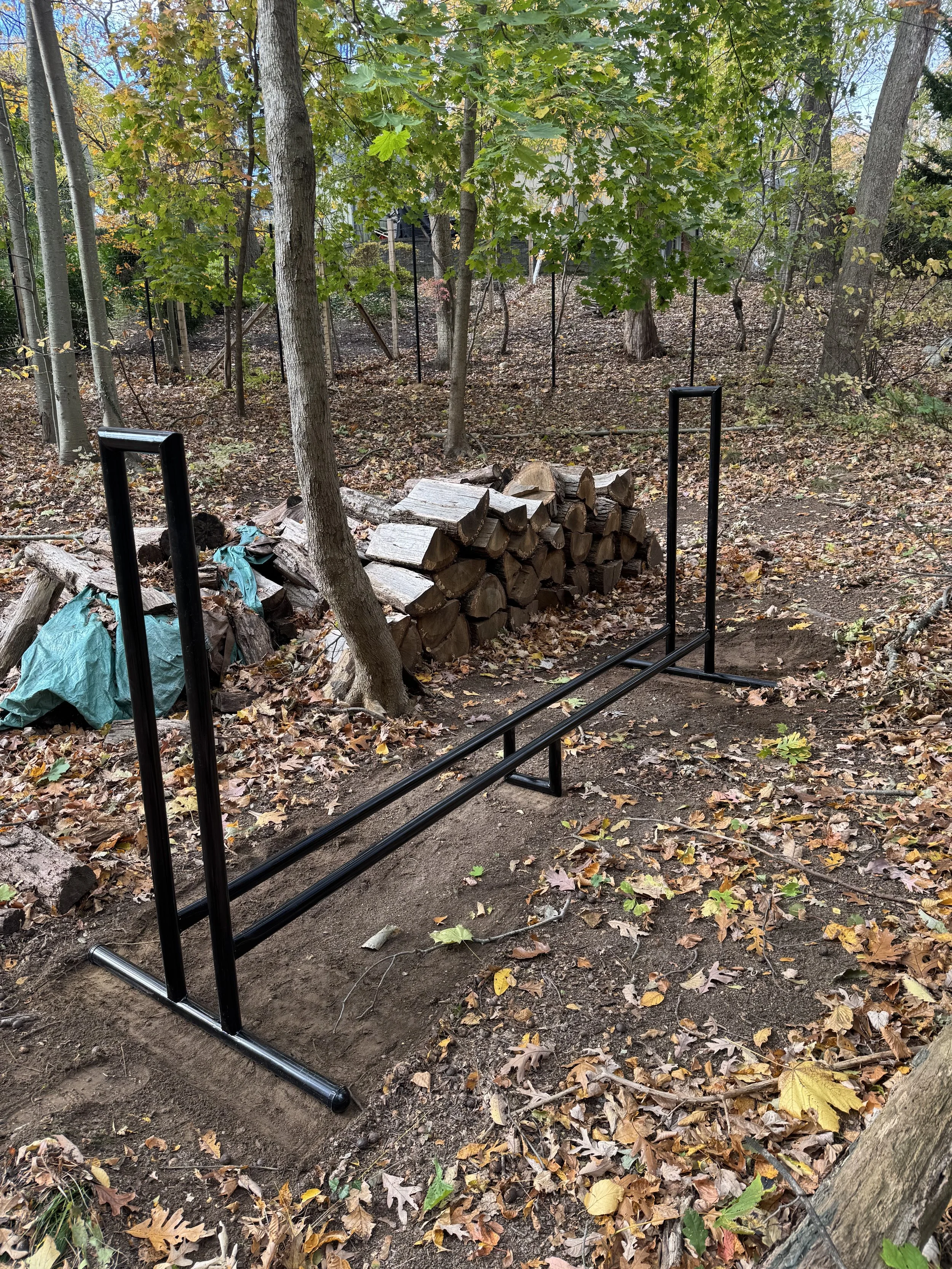A partially assembled black metal garden bed frame set up on bare soil among fallen autumn leaves in a wooded backyard with trees and a pile of chopped firewood.