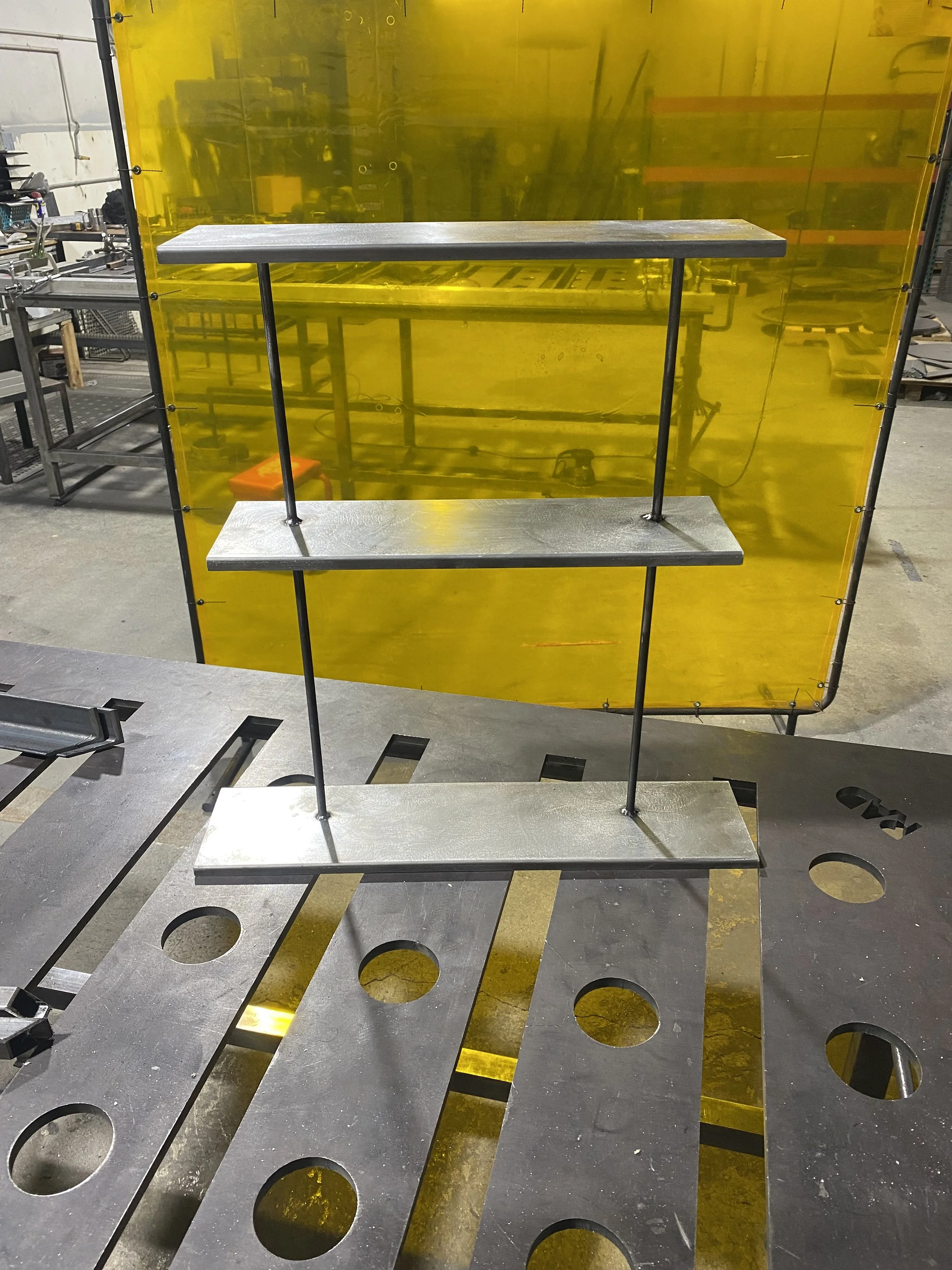 Metal shelving unit with three horizontal shelves, supported by two vertical rods, on a workshop table with various tools and equipment in the background.