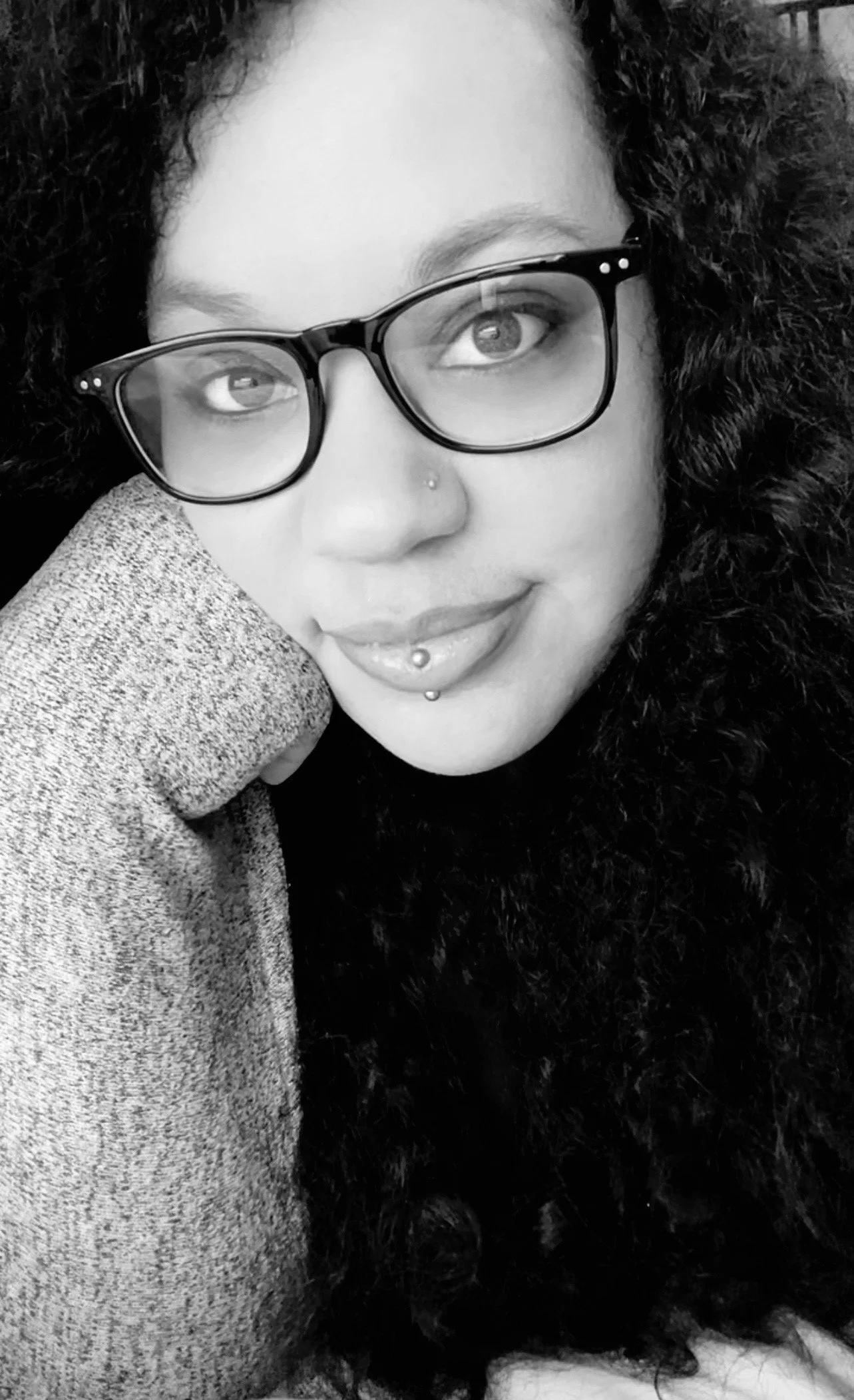 A woman with glasses and curly hair, smiling, with a piercing on her lower lip.