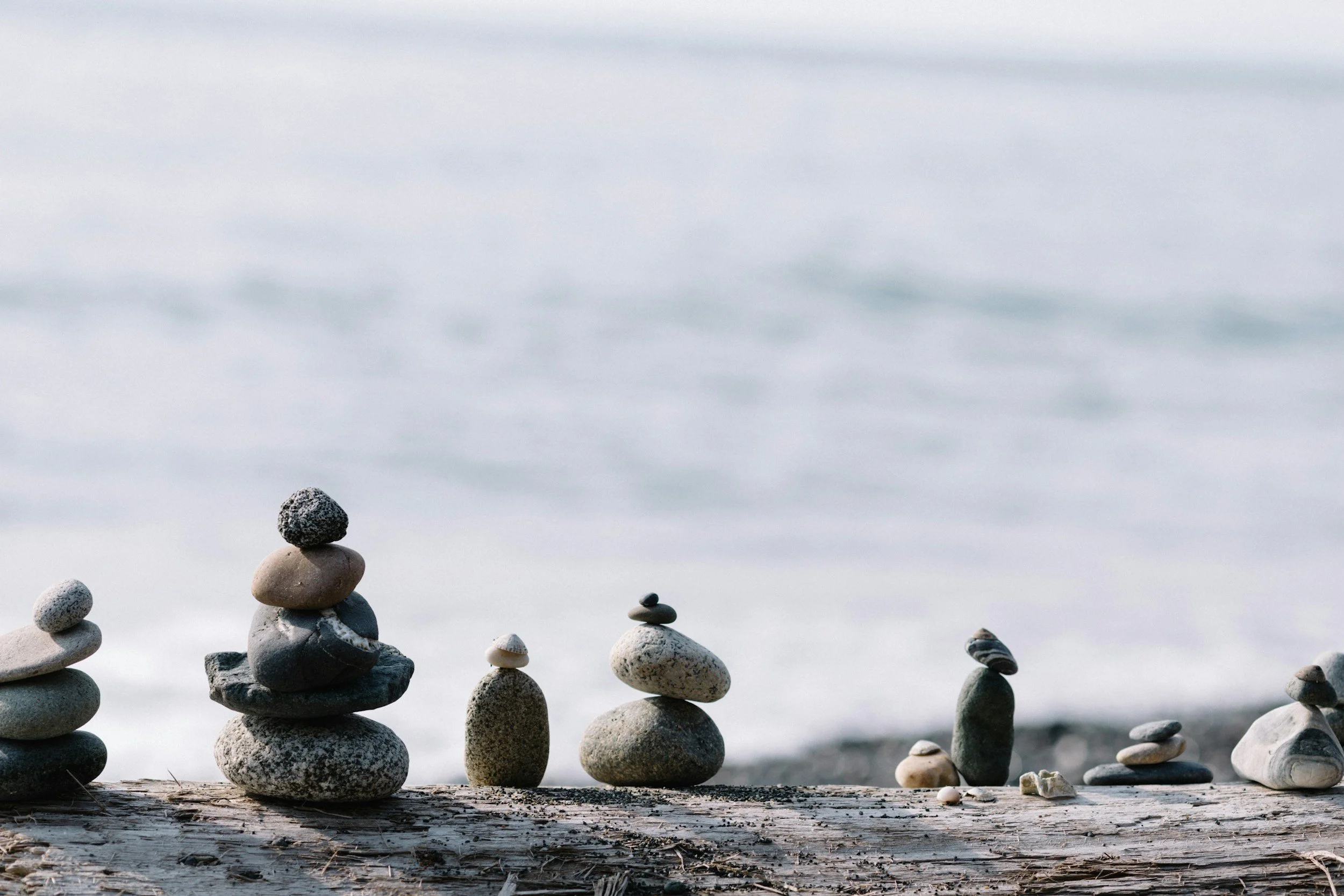 Multiple stacked rocks and pebbles on a weathered wooden log near the water, with gentle waves in the background.