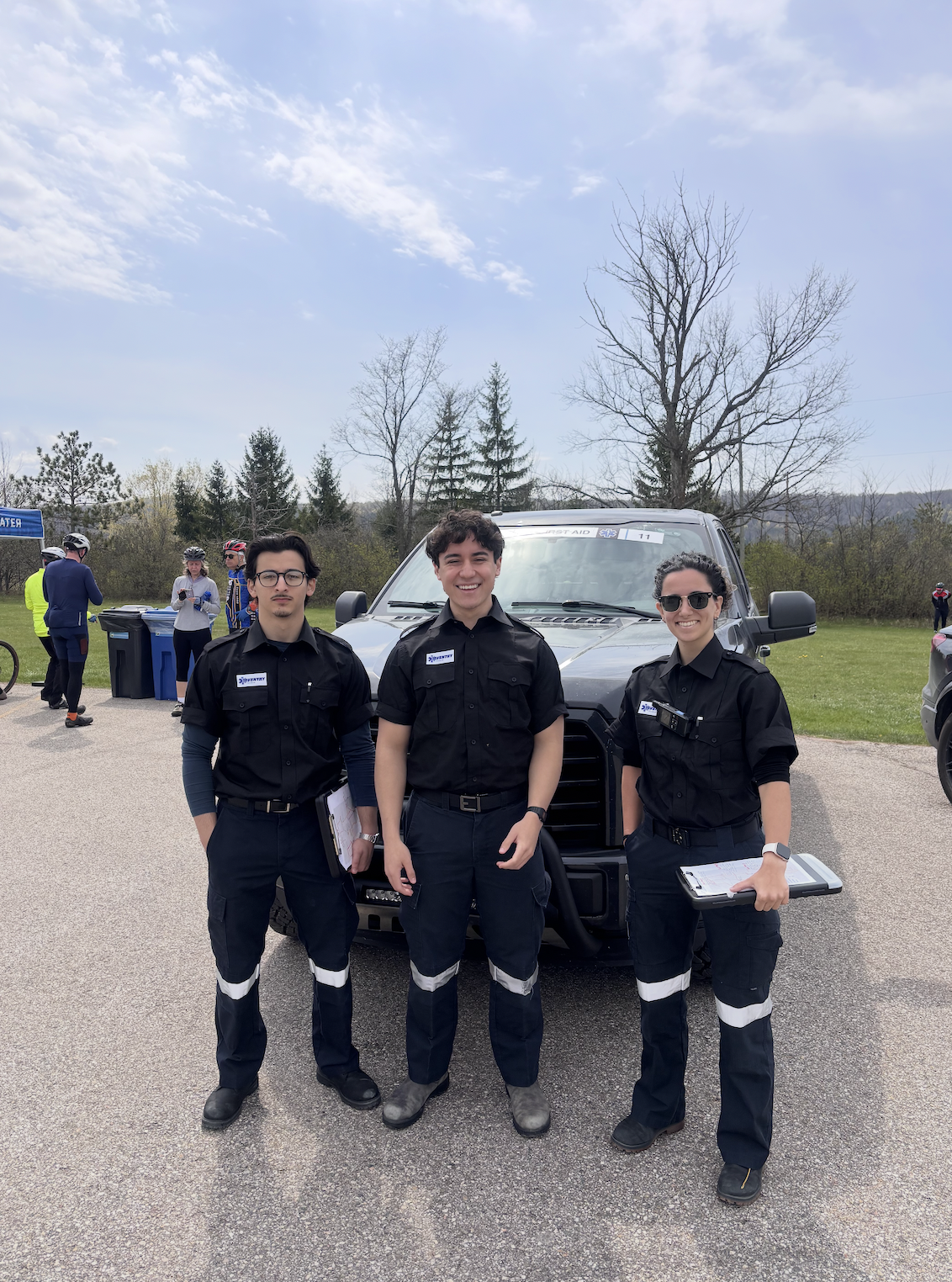 Three Aventry emergency responders in black uniforms standing in front of a black vehicle during an outdoor event, with other people and trees in the background.