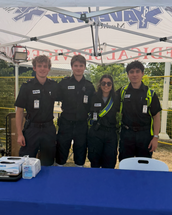Four young uniformed emergency responders, three males and one female, standing together under a white canopy with emergency medical symbols, outside at a fenced area with trees in the background.