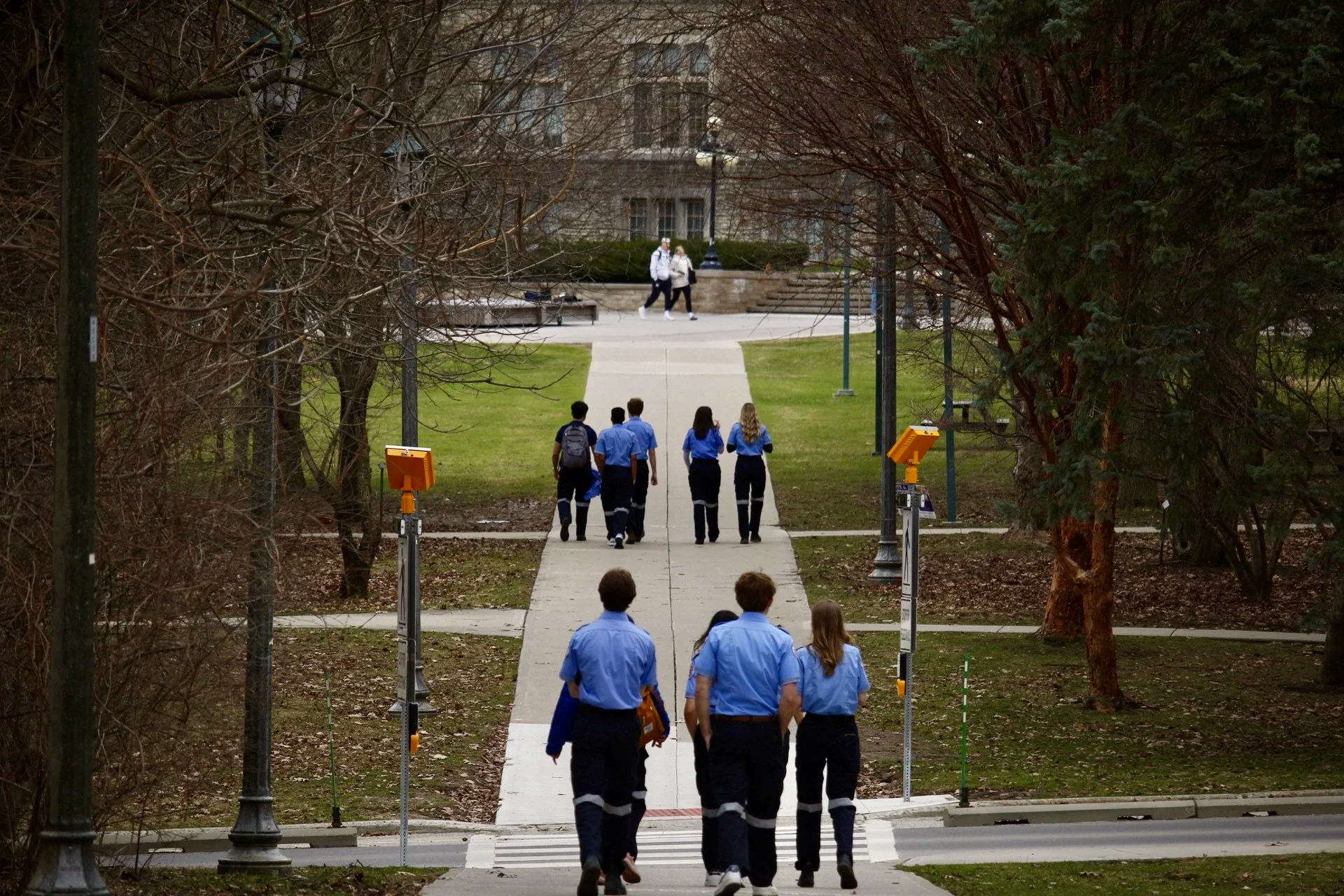 Group of five students in blue uniforms walking on sidewalk through park, with a larger group of students ahead and two people walking on the path in the distance.