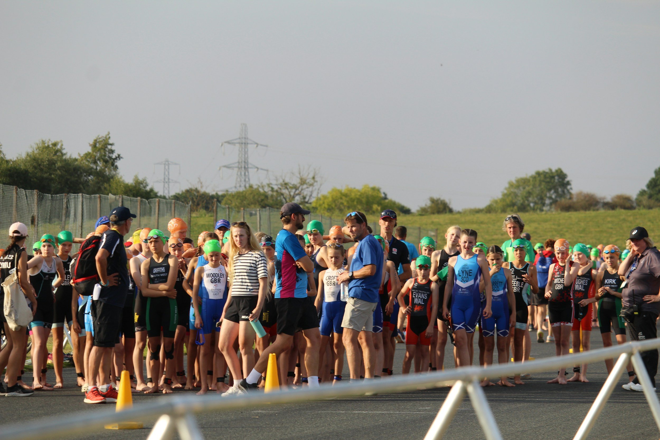 Swimmers gather at the starting line in a triathlon, preparing for the race with coaches giving instructions on a sunny day.