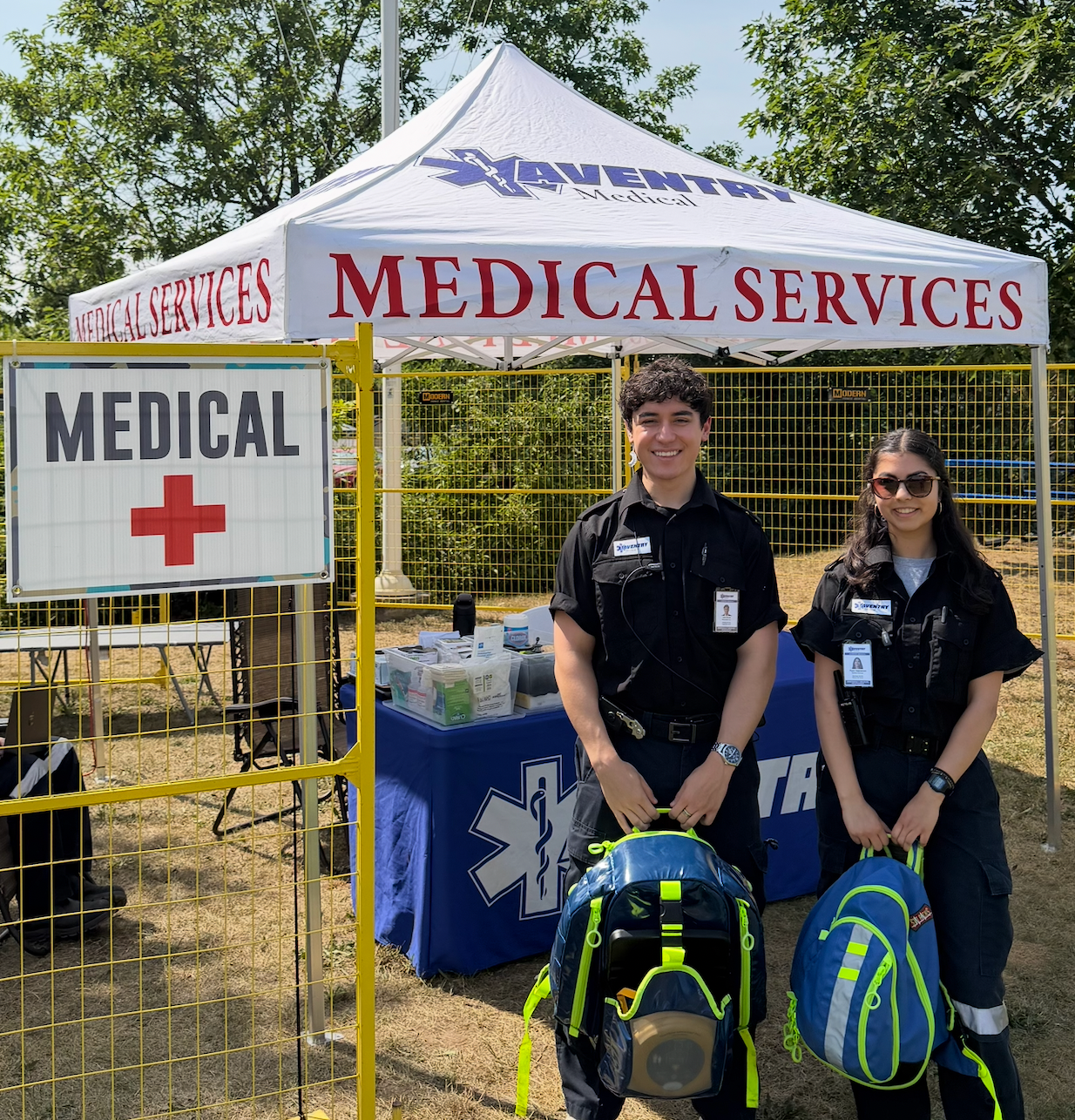 Two emergency medical technicians standing in front of a medical tent labeled 'Medical Services' at an outdoor event. They are holding backpacks and smiling, wearing black uniforms with emergency IDs, near a table with medical supplies.