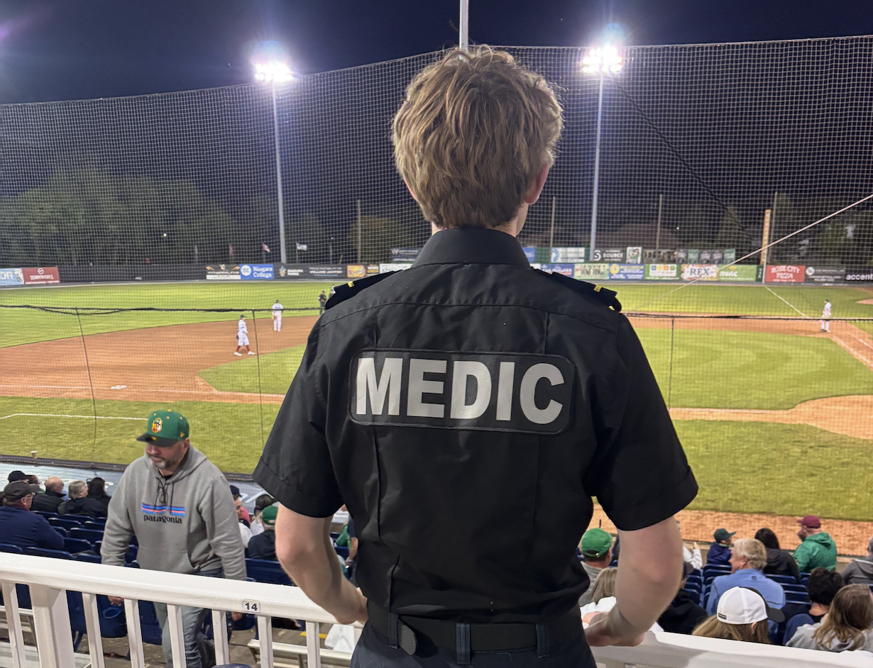 A person wearing a black rescue medic shirt watching a baseball game from the stands at night. The baseball field is lit up, with players on the field and spectators seated.