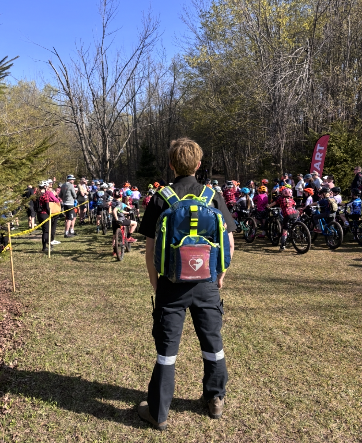 A young Aventry medic with a blue backpack standing in front of a large group of children on bicycles, all wearing helmets at an outdoor event in a wooded area.
