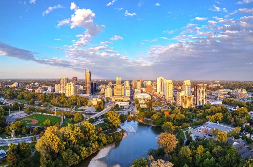 Aerial view of a cityscape with tall buildings, a river, a park with a baseball field, and a partly cloudy sky.