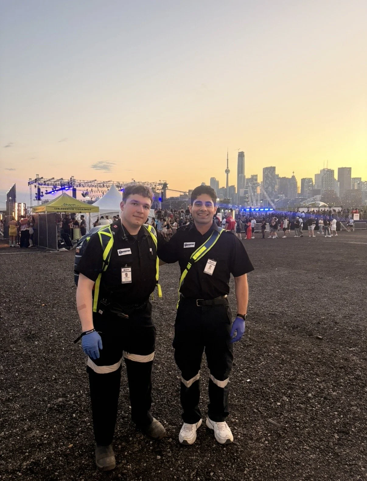 Two emergency responders in black uniforms with yellow reflective straps and gloves stand outside during sunset at a crowded event. The city skyline with tall buildings and a tower can be seen in the background, along with a stage and tents.