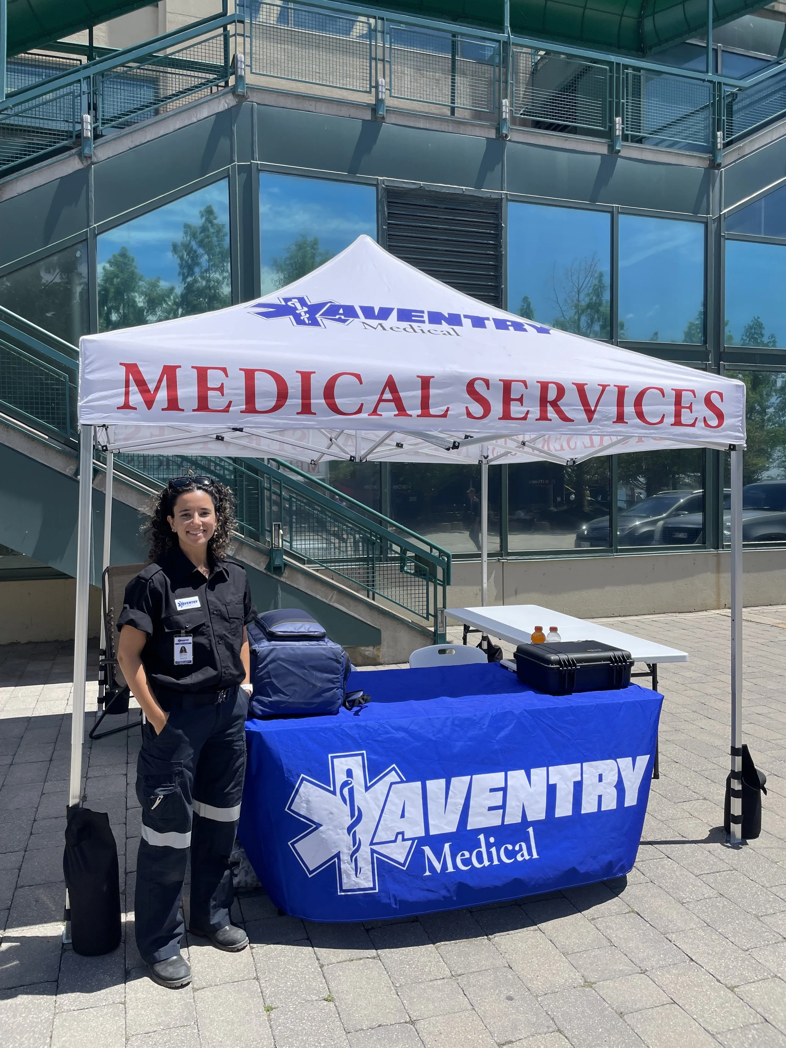 A woman in a paramedic uniform standing under a tent with 'Medical Services' and 'Aventry' logos. The tent is set up outside a glass building with a staircase, and there are medical supplies on the table in front of her.