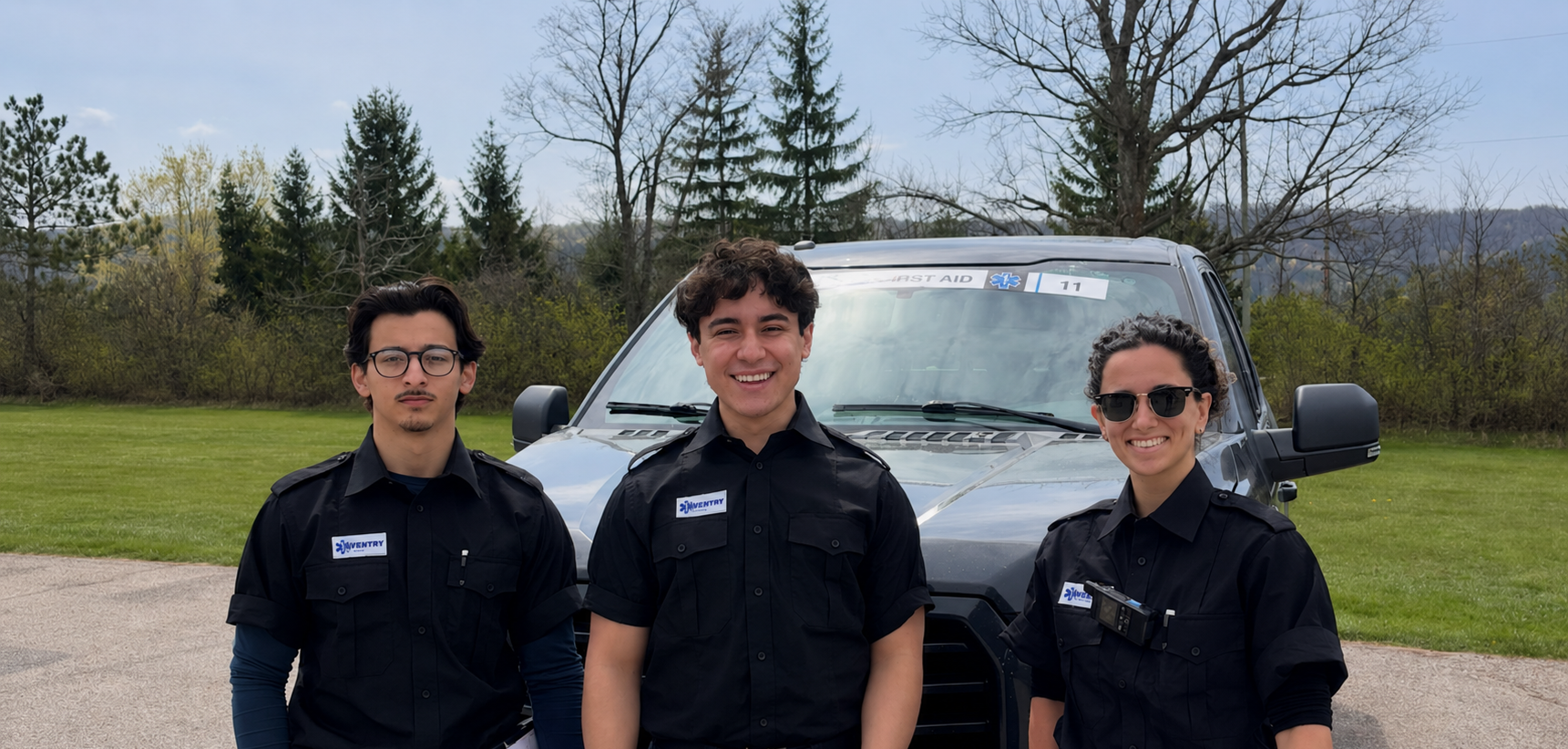 Three Aventry responders standing in front of a gray emergency vehicle outdoors, with trees and a cloudy sky in the background.