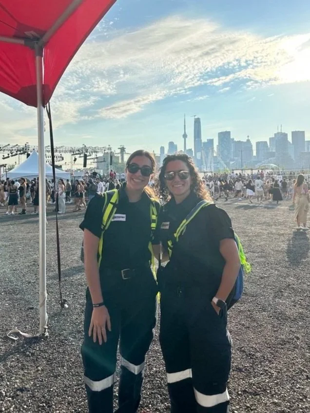 Two Aventry medics in black uniforms and sunglasses standing outdoors at a crowded event in front of a city skyline with tall buildings.