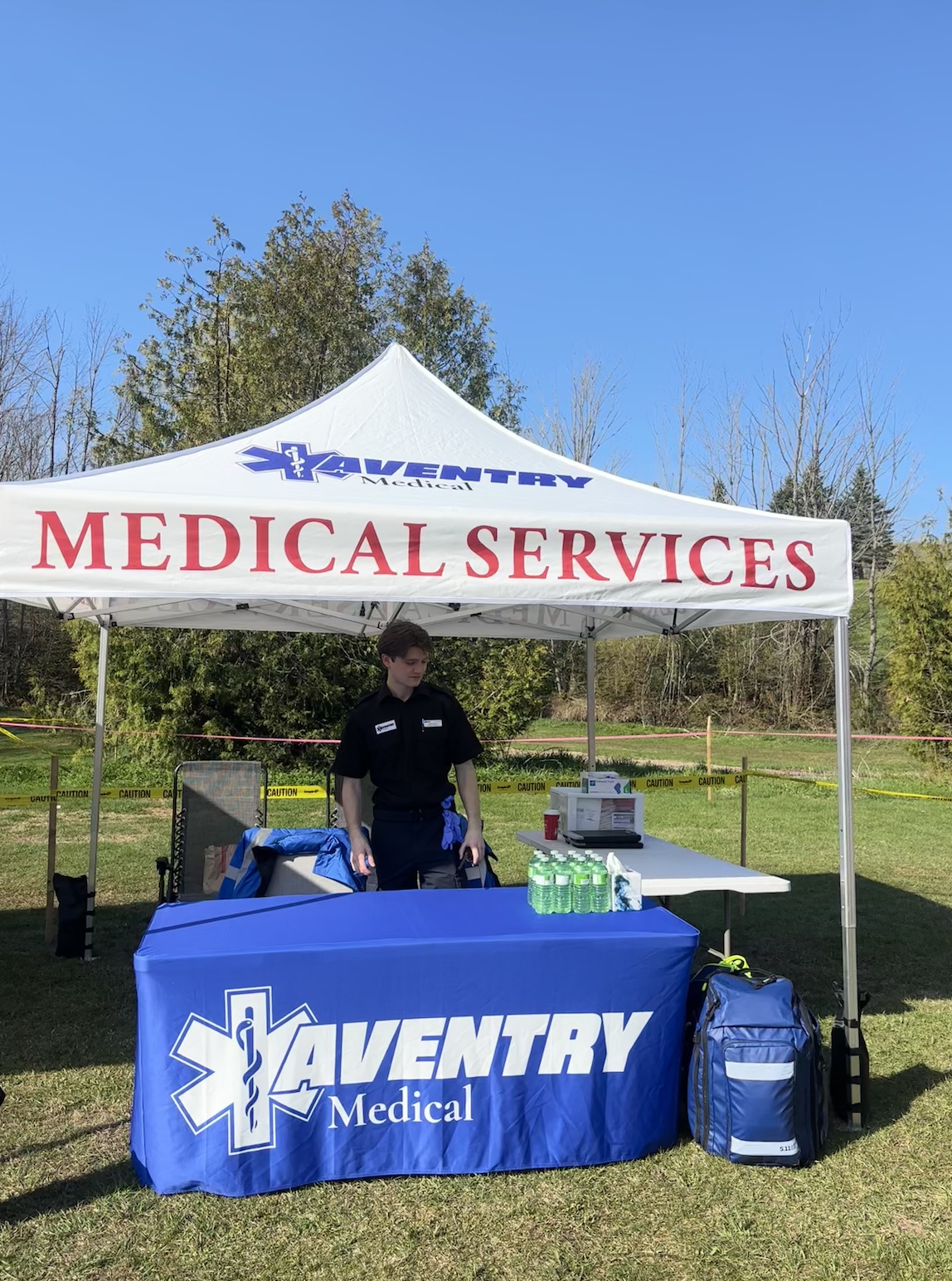 A paramedic standing under a white tent with signs that read 'Medical Services' and 'AVENTRY Medical.' The tent is set up outdoors on grass with trees in the background. There are bottled waters, a first aid kit, and supplies on tables, and caution tape marks off an area behind the tent.