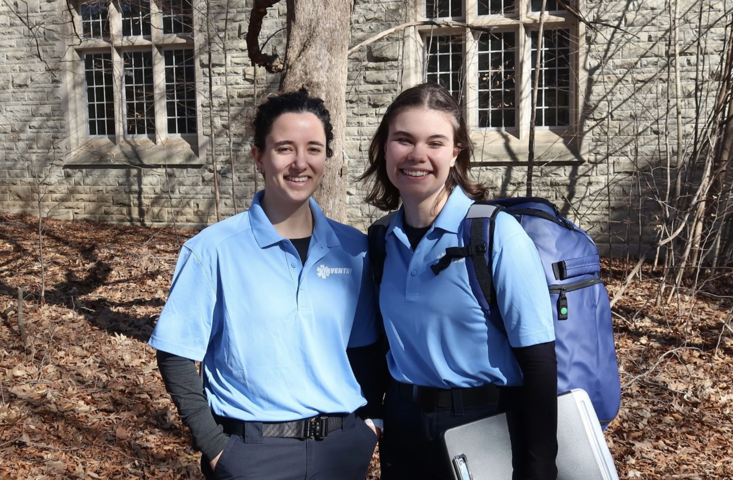 Two young Aventry Medical women smiling outdoors in front of a stone building and leafless trees, wearing blue uniforms with one carrying a backpack and a plastic container.
