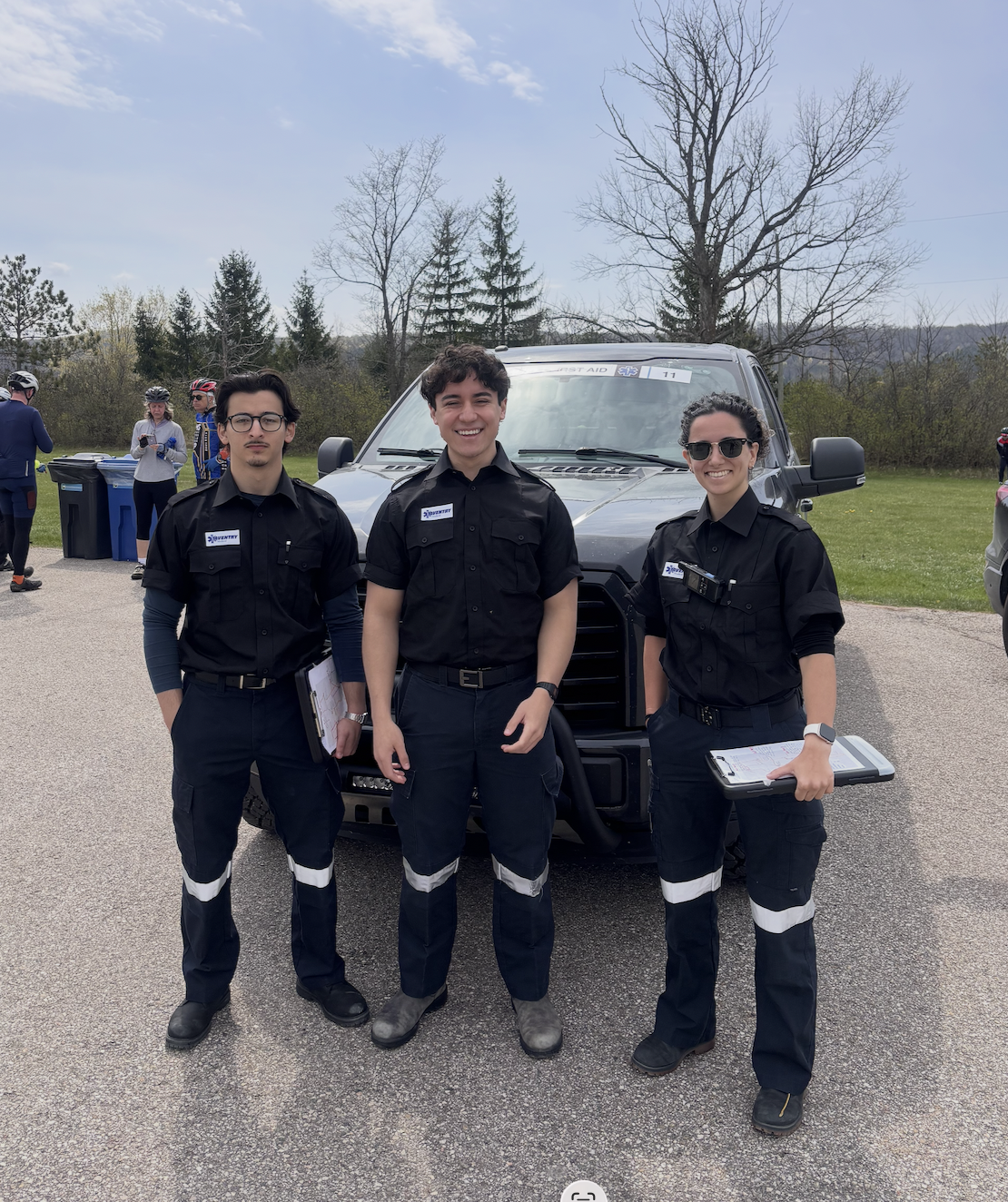 Three Aventry emergency responders in black uniforms standing in front of a gray emergency vehicle outdoors with trees and a few people in the background.